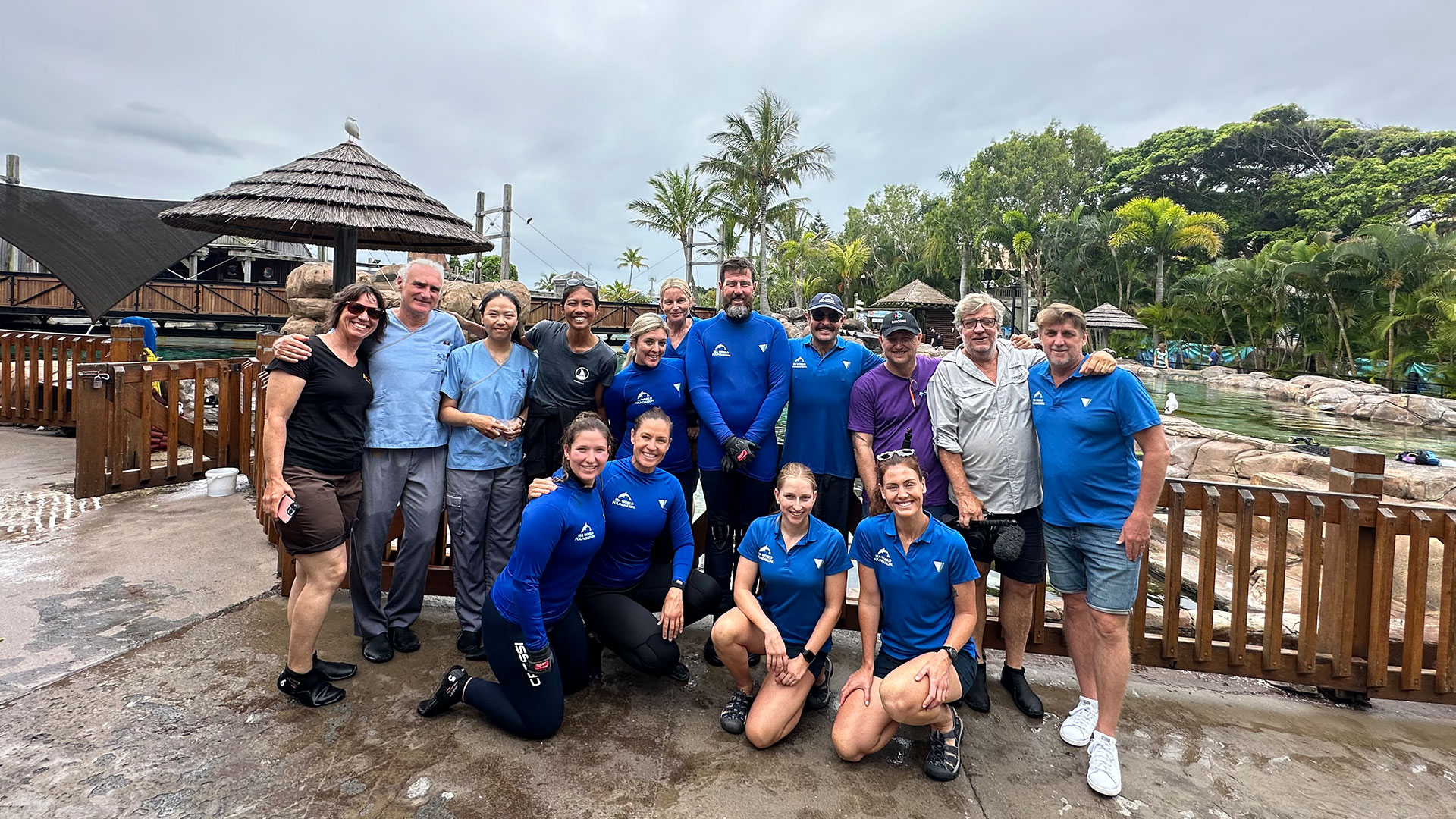 A group of people, including some in blue wetsuits, pose together outdoors near a fenced area with tropical plants and thatched structures.