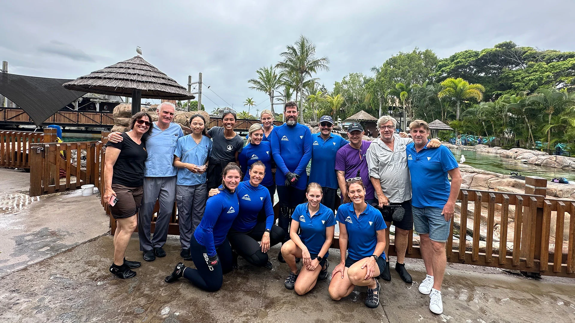A group of people, including some in blue wetsuits, pose together outdoors near a fenced area with tropical plants and thatched structures.