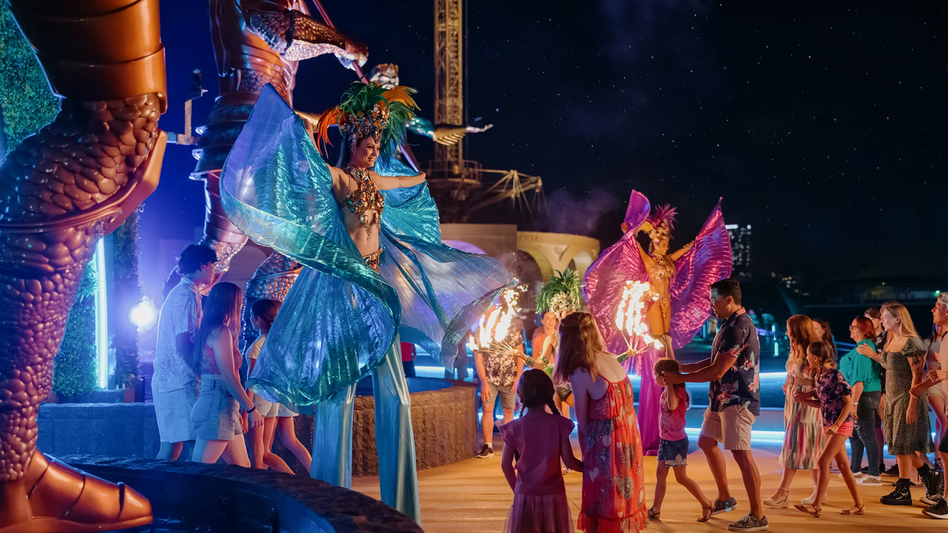Performers in colorful costumes entertain a crowd of adults and children at night, with bright lights and a festive atmosphere. Some performers are on stilts and others hold props, while families watch and interact.