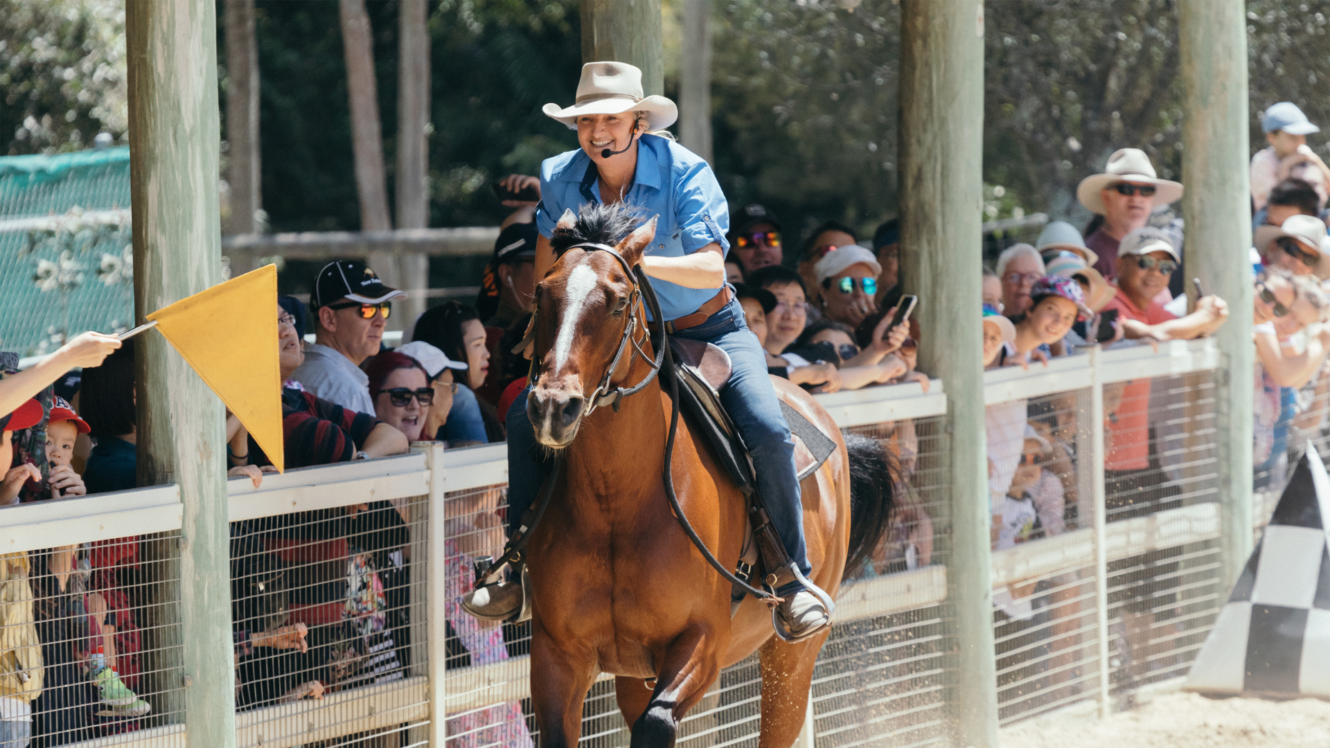 A person wearing a blue shirt and hat rides a horse on a dirt track, lined with spectators behind a fence, during a show at Paradise Country.