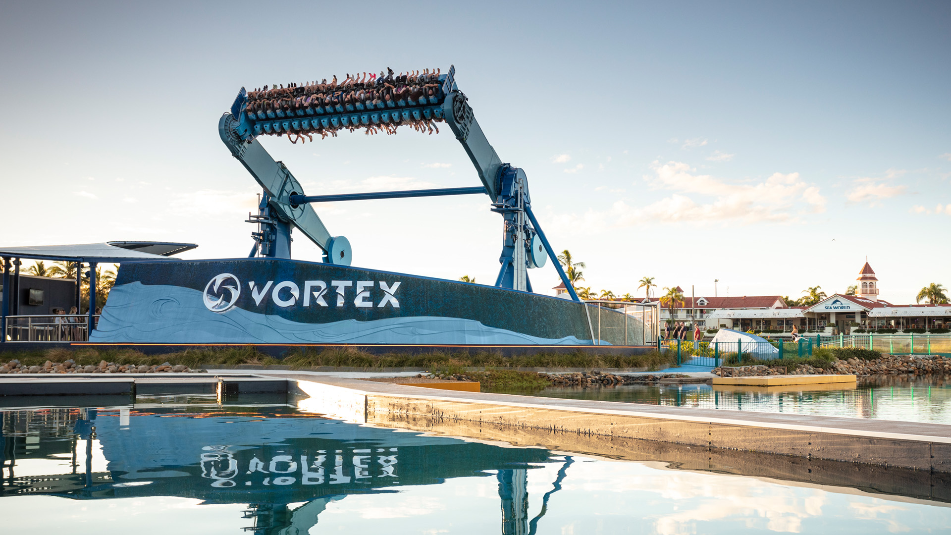 Large amusement park ride labeled "Vortex" flips riders upside down above a pool of water, with blue sky and park buildings in the background.