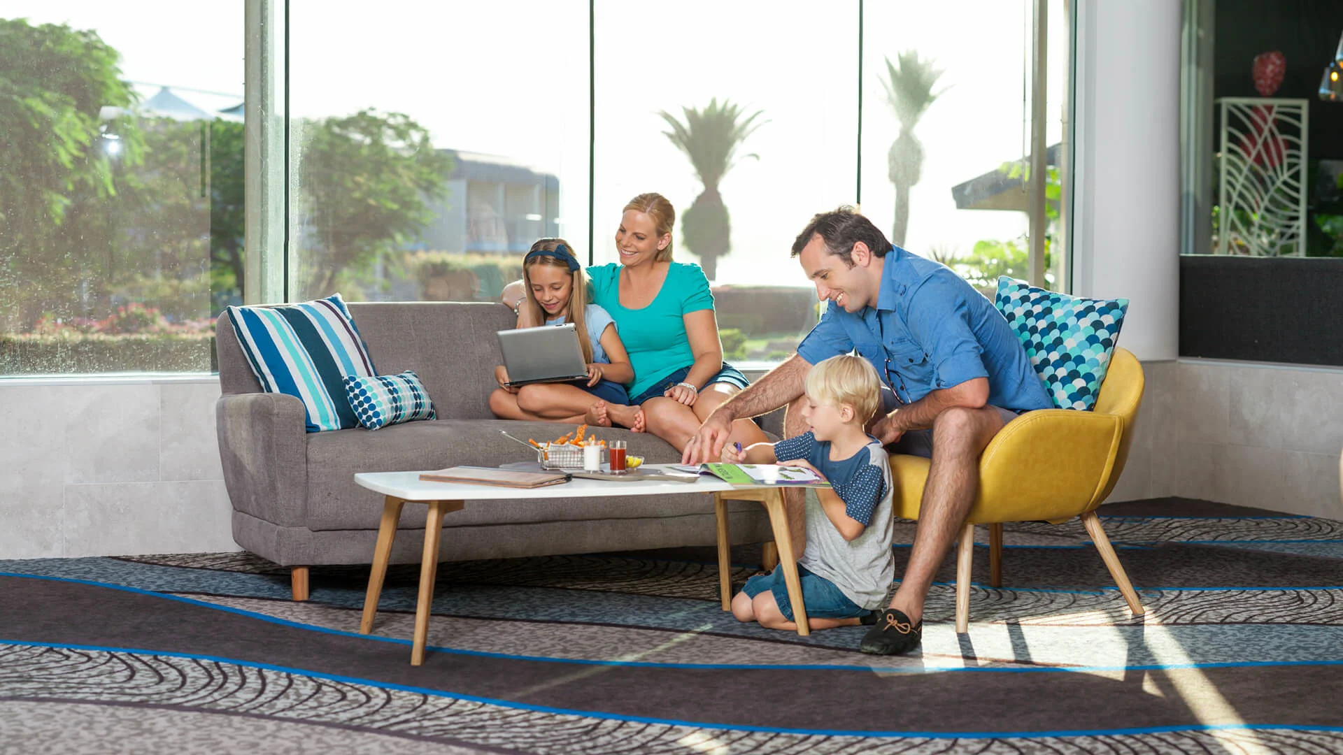 A family of four enjoys indoor activities in a bright living room. Two adults sit on a sofa, while two children engage with a coffee table. Large windows reveal trees outside.