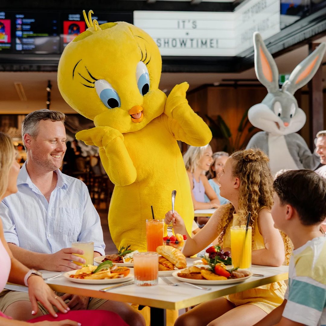 A family enjoys a meal at a restaurant as a person in a large Tweety Bird costume interacts with them. Bugs Bunny stands in the background. Plates of food and drinks are on the table.