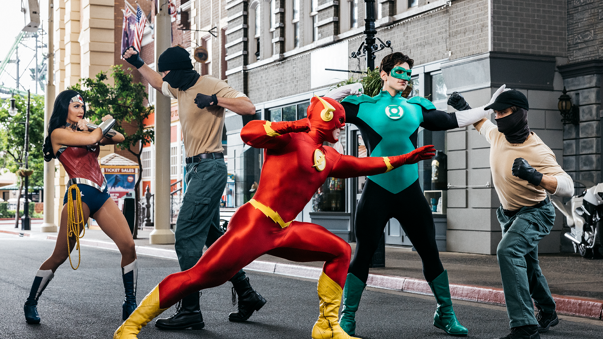 Four people dressed as superheroes are posing in a staged combat scene with two individuals wearing masks on a city street. Wonder Woman flash Green Lantern