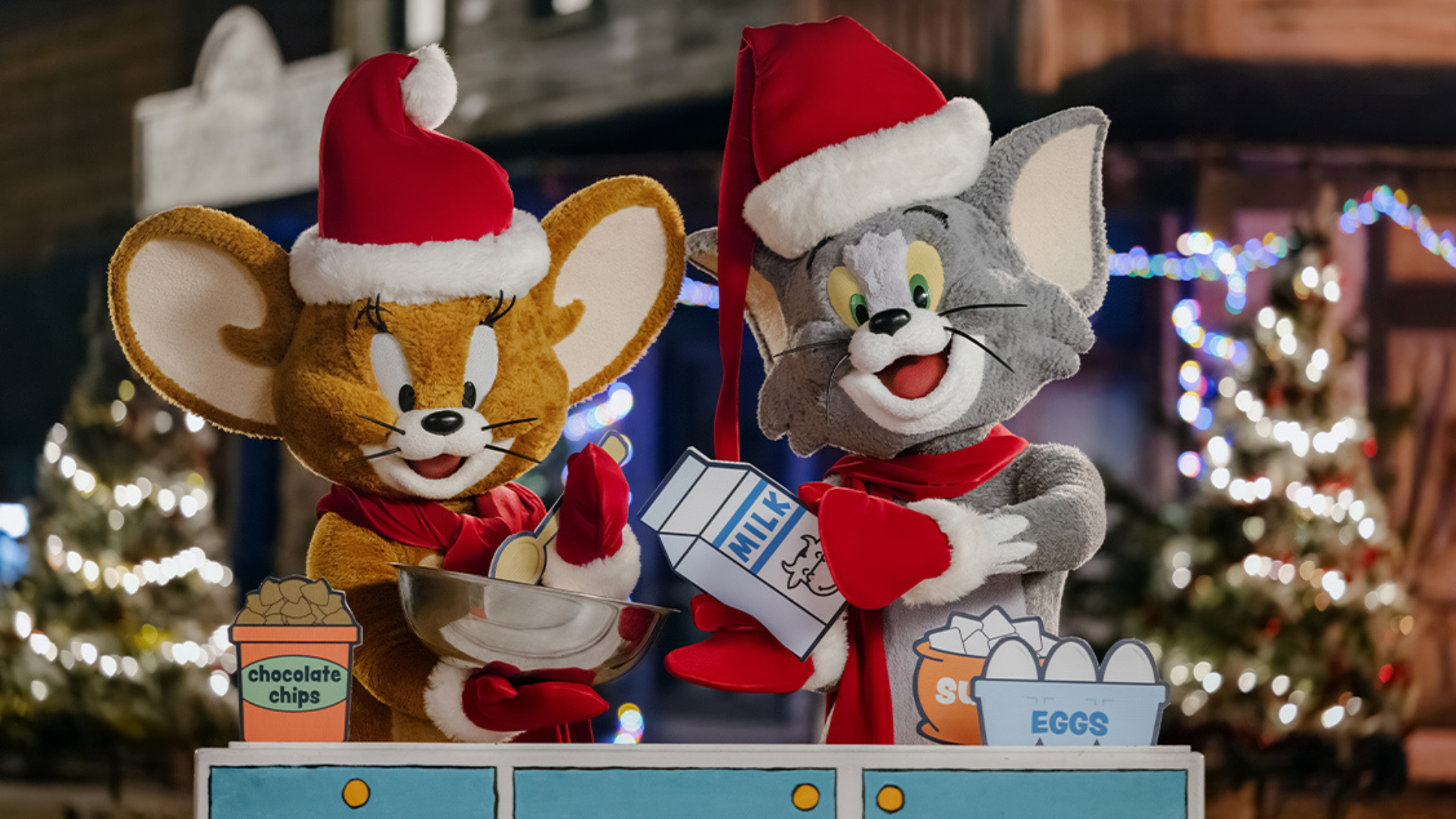 Jerry and Tom, dressed in Santa hats and red scarves, prepare holiday cookies together with ingredients like chocolate chips, eggs, and milk. Festive lights and decorated trees are visible in the background.