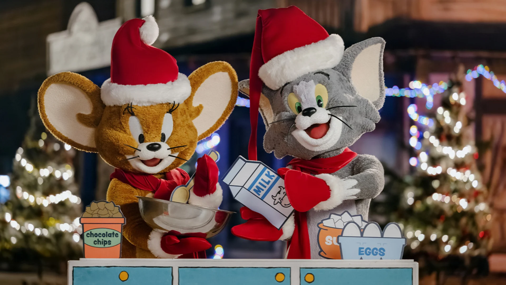 Jerry and Tom, dressed in Santa hats and red scarves, prepare holiday cookies together with ingredients like chocolate chips, eggs, and milk. Festive lights and decorated trees are visible in the background.