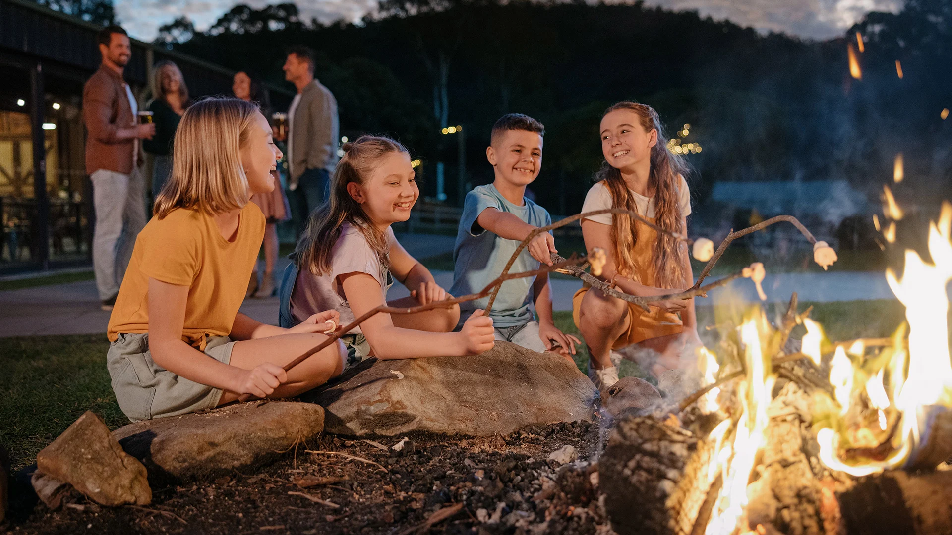 Four children roast marshmallows over a campfire, with adults talking in the background.