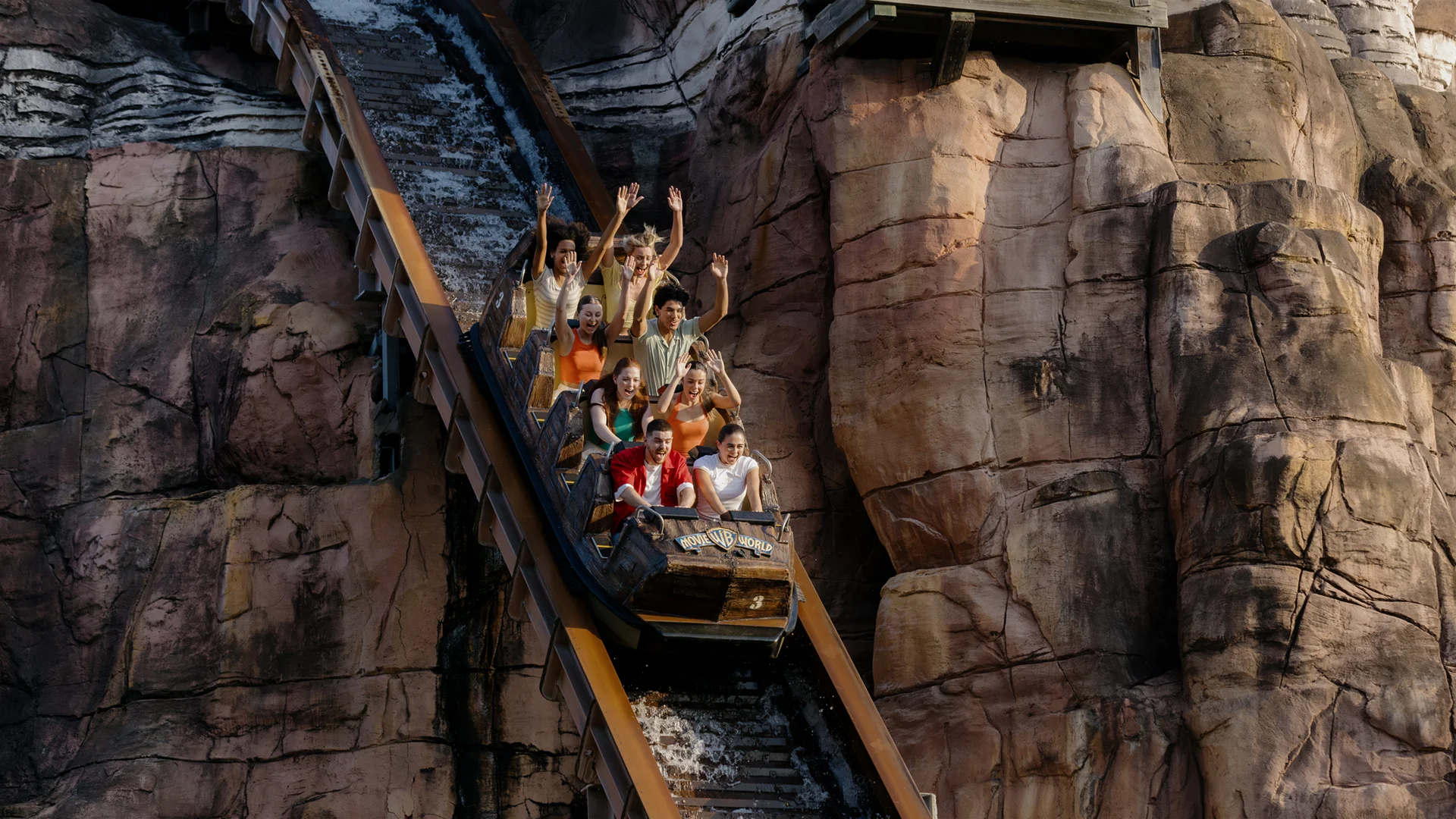 A group of people ride a steep roller coaster down a mountain-themed track, some with arms raised in excitement, surrounded by rugged, rocky scenery.