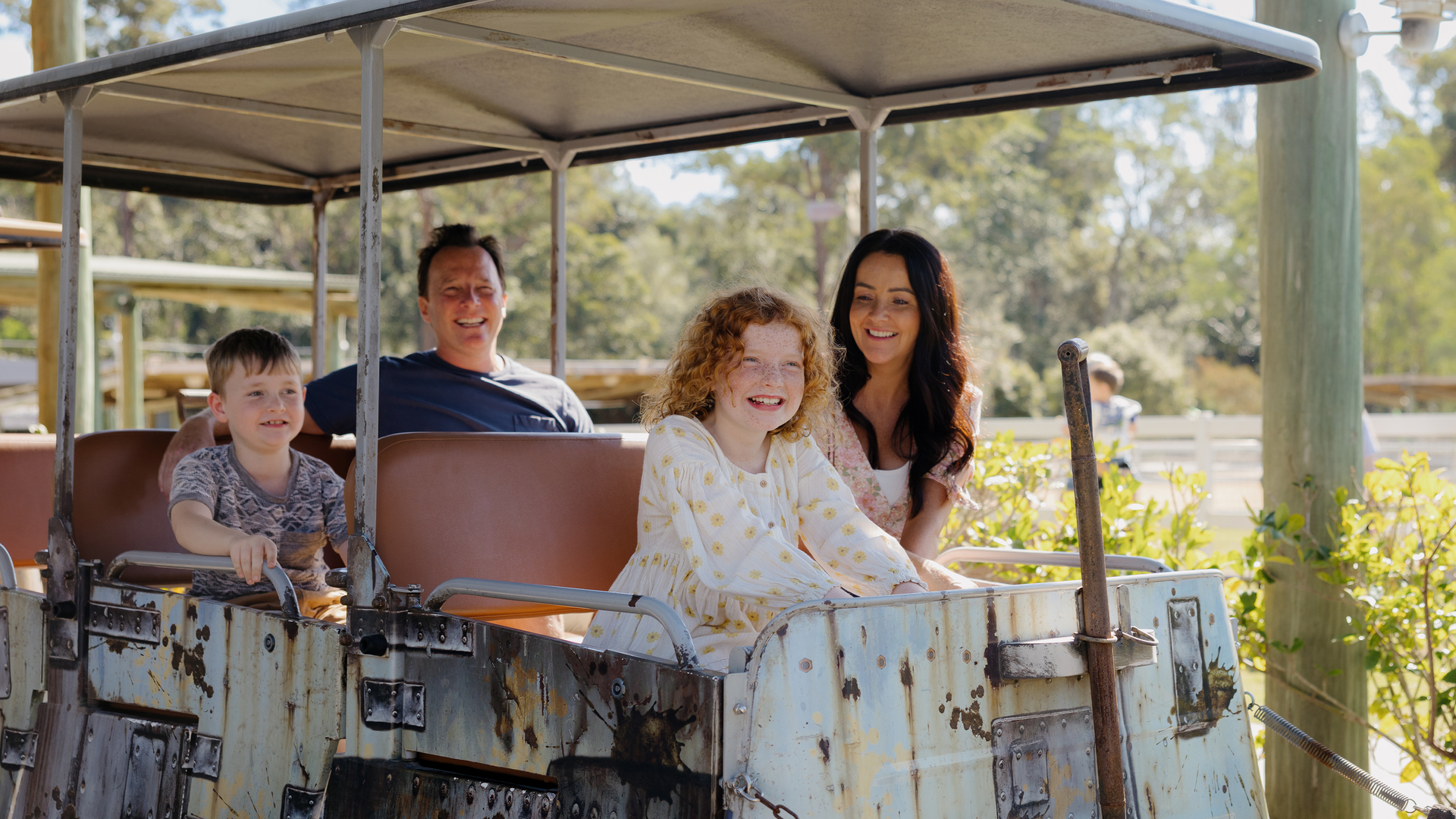 A smiling family of four—two adults and two children—sit together in an old, open-air amusement park ride on a sunny day, surrounded by greenery.