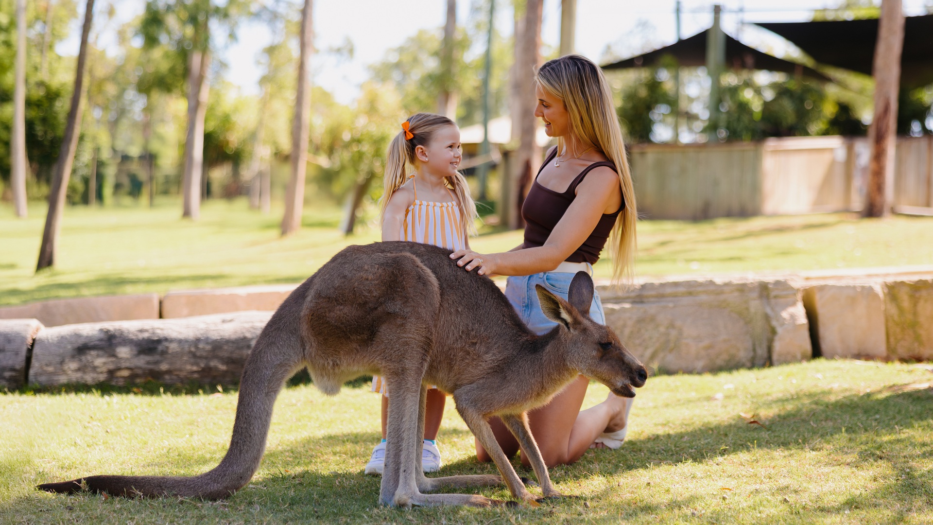 A woman and a young girl smile and kneel on the grass while petting a kangaroo in a sunlit outdoor park.