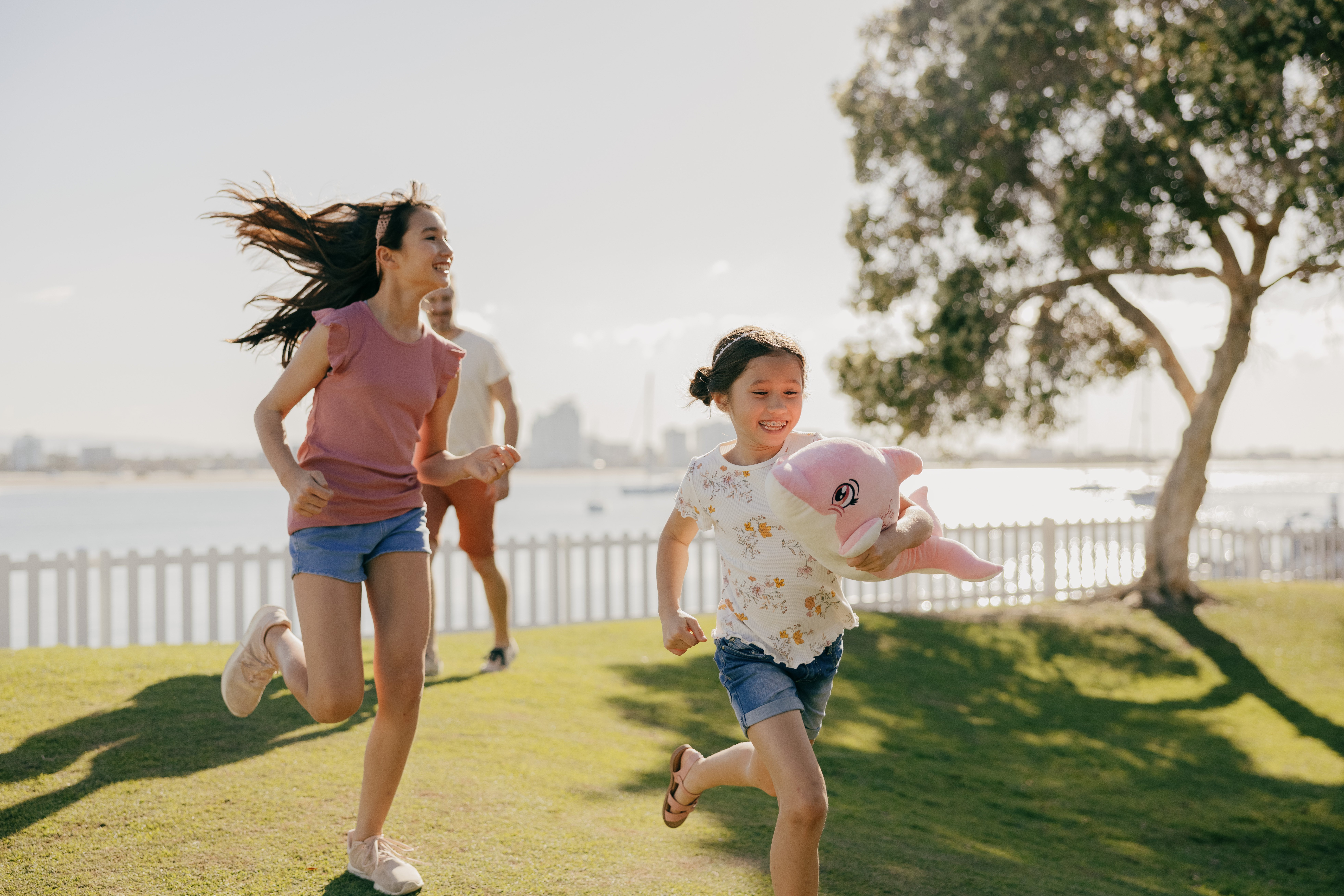 Two children run and play on grass near a body of water; one holds a pink stuffed toy. A tree and white fence are in the background on a sunny day.