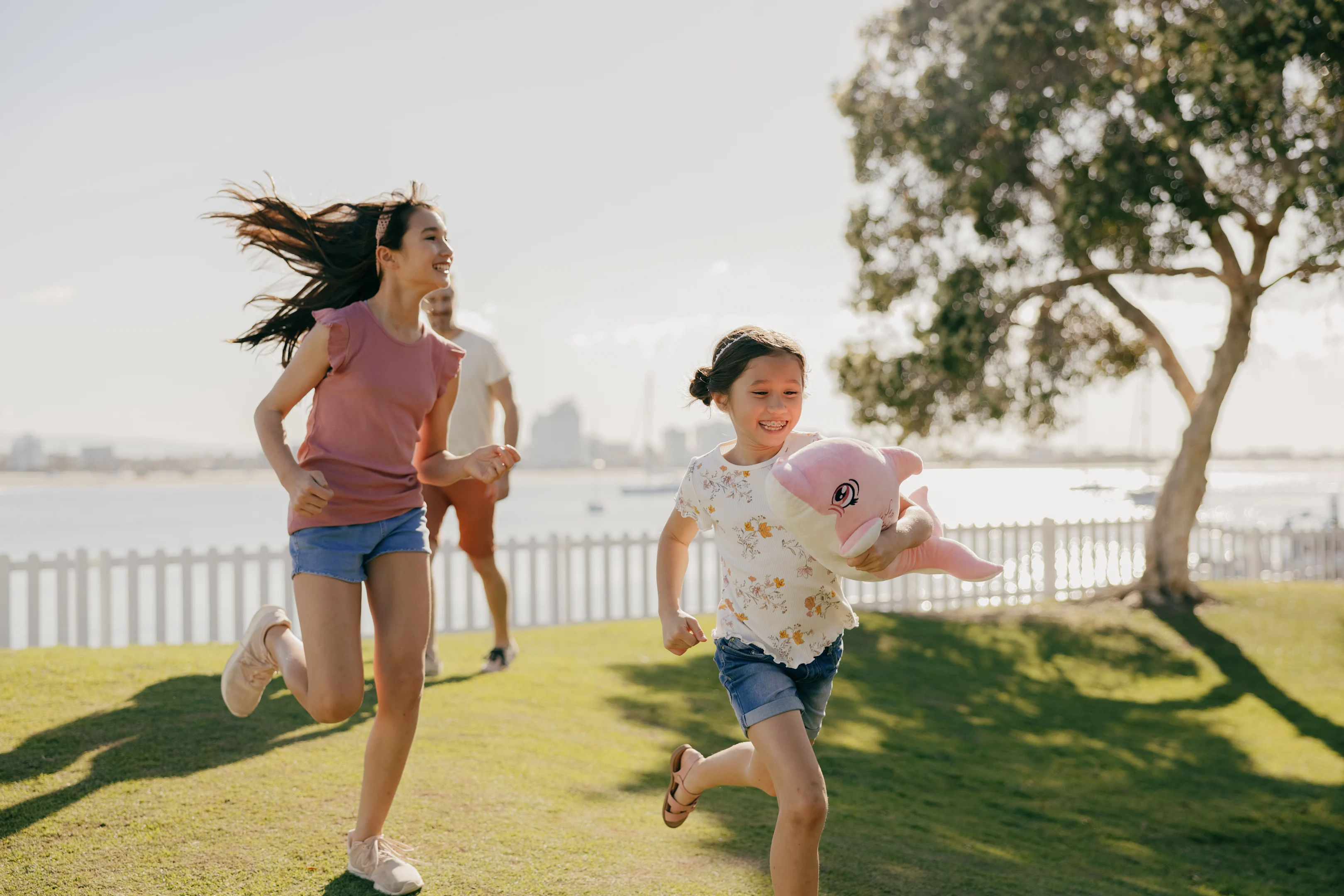 Two children run and play on grass near a body of water; one holds a pink stuffed toy. A tree and white fence are in the background on a sunny day.