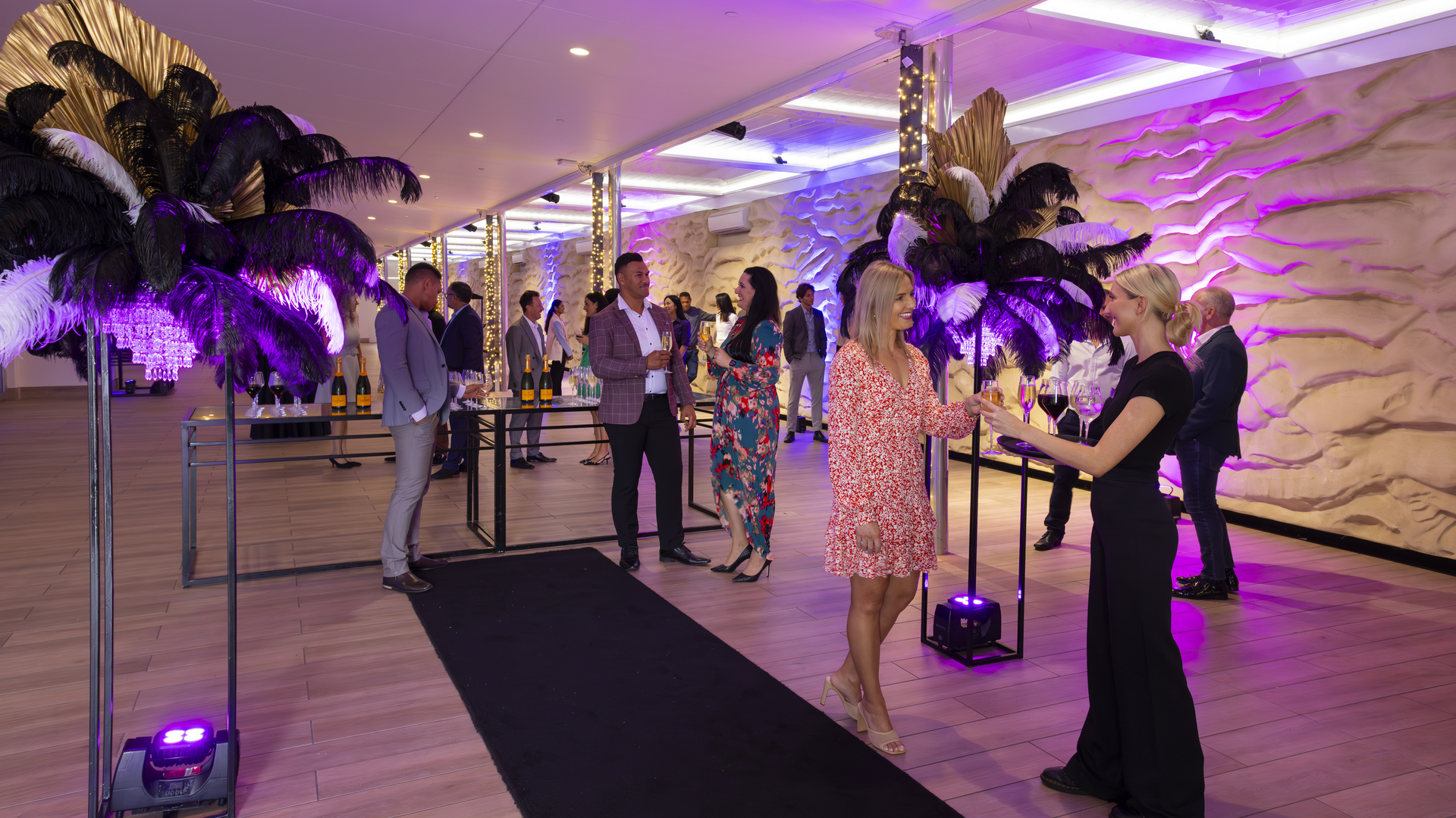 People dressed in semi-formal attire mingle at a stylish indoor event. Decorative feather arrangements and purple lighting enhance the upscale atmosphere, while drinks are served near a textured, wavy wall.