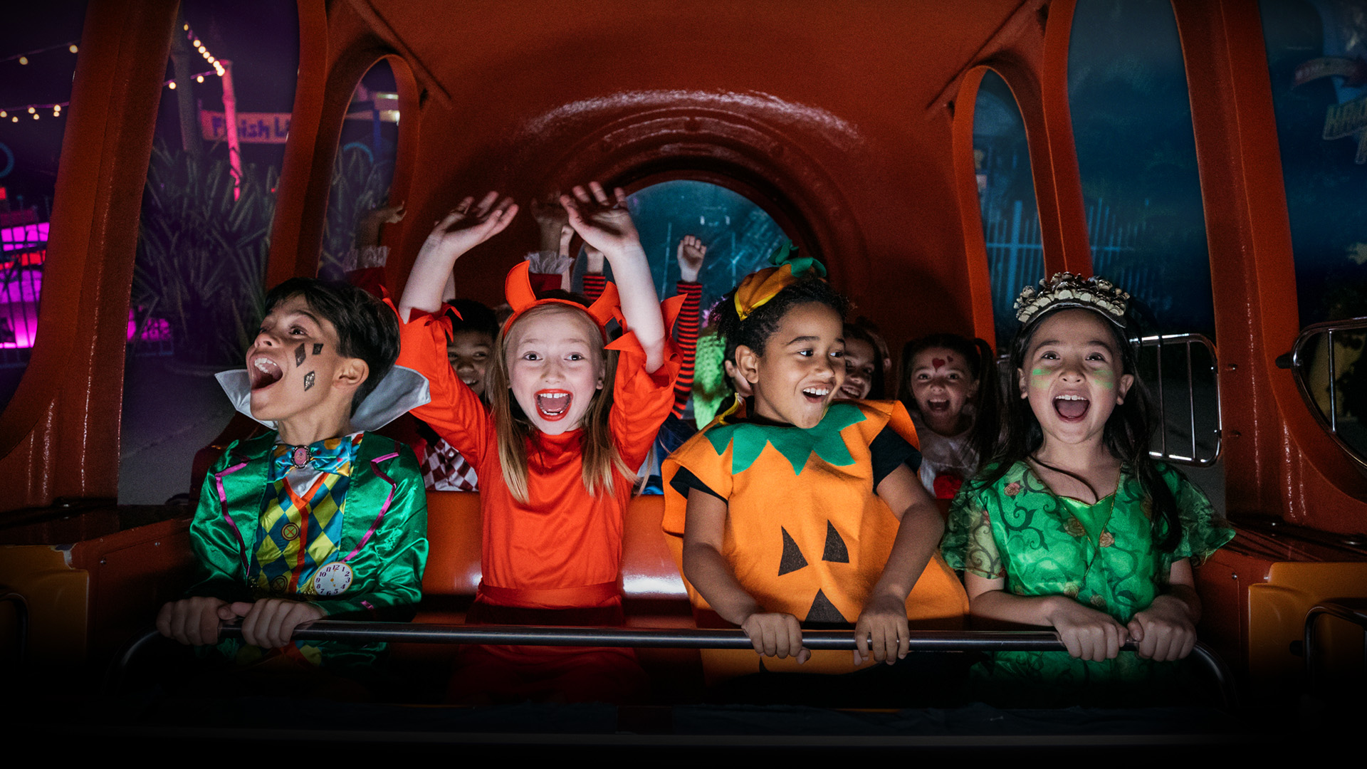 A group of children in colorful Halloween costumes excitedly ride an amusement park attraction, smiling and raising their hands in delight under vibrant lights.