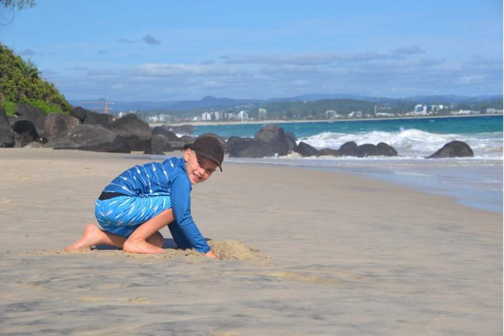 A young boy in a blue rash guard and swim trunks crouches and plays in the sand on a sunny beach, with waves, rocks, and distant buildings visible in the background.