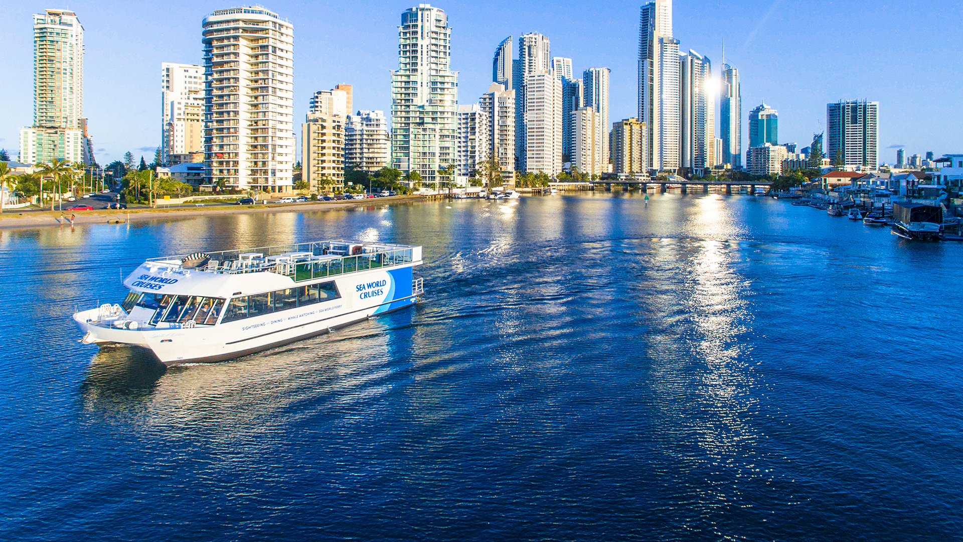A white sightseeing boat cruises on calm blue water near a city skyline with tall modern buildings under a clear sky, reflecting in the water.