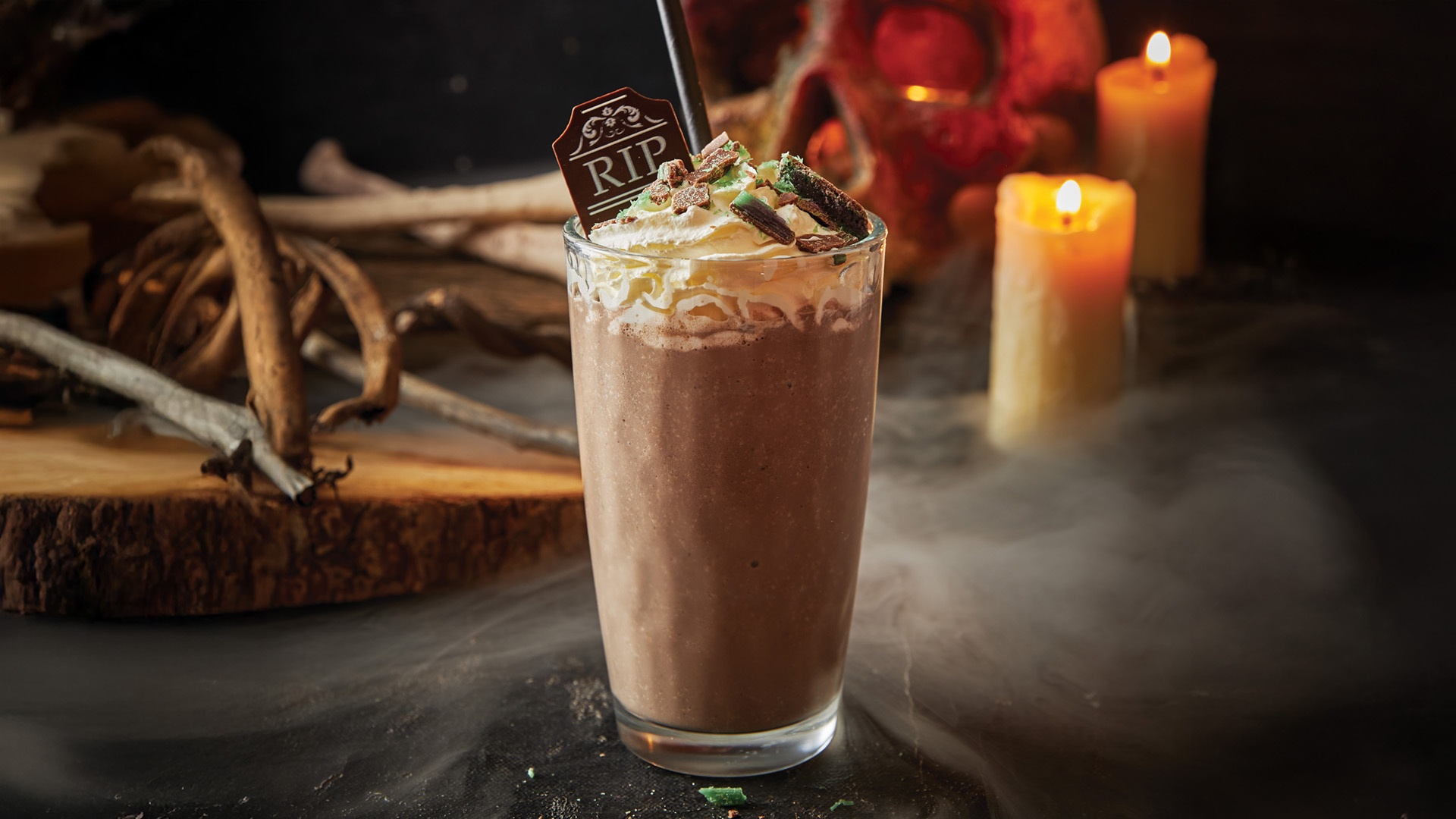A chocolate milkshake topped with whipped cream, chocolate pieces, and a "RIP" tombstone decoration, sits on a smoky, dark surface with candles and bones in the background, creating a spooky Halloween vibe.