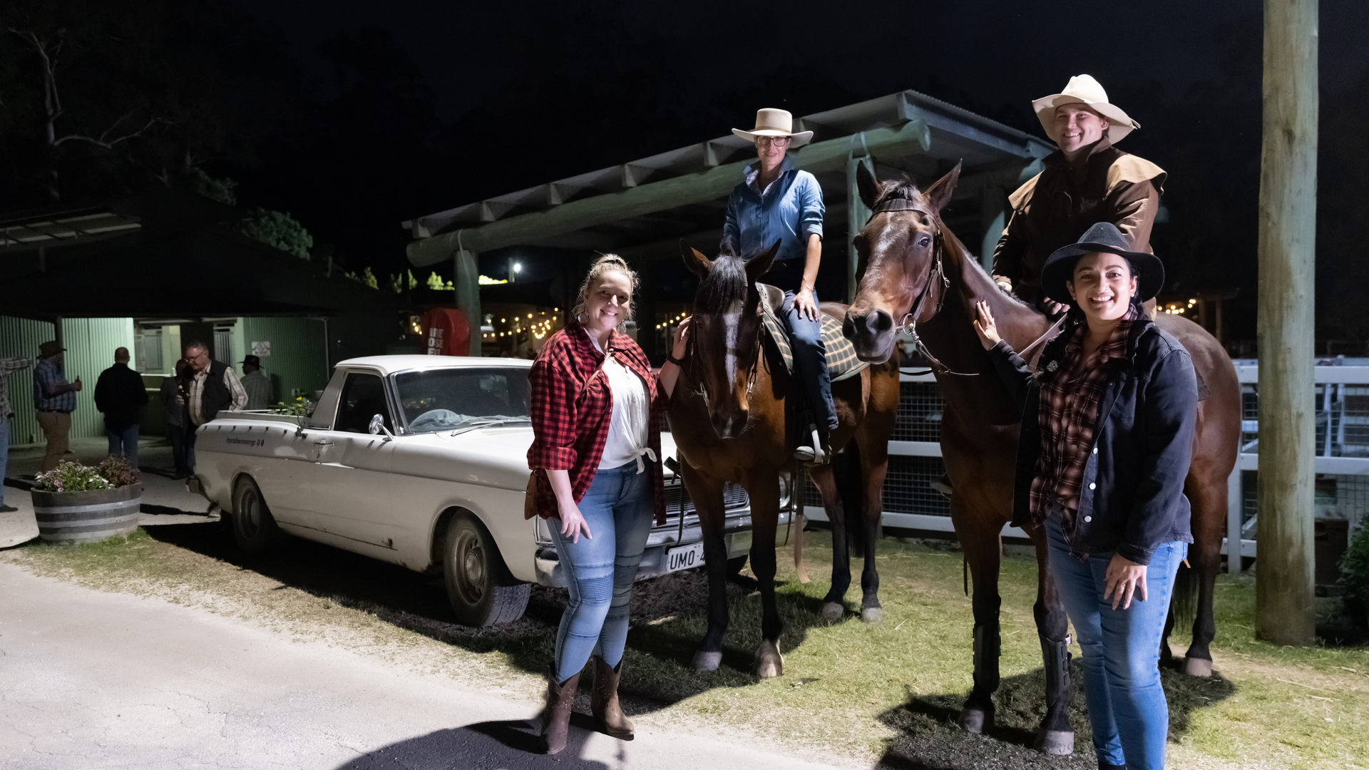 Four people pose at night; two are standing near a white classic car, and two are on horseback wearing cowboy hats and western attire.