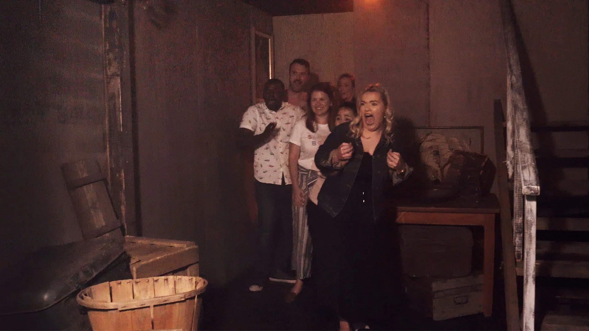 A group of six people stand huddled together in a dimly lit, rustic room. One woman in front appears startled, with her mouth open and hands raised, while the others look on nervously behind her.