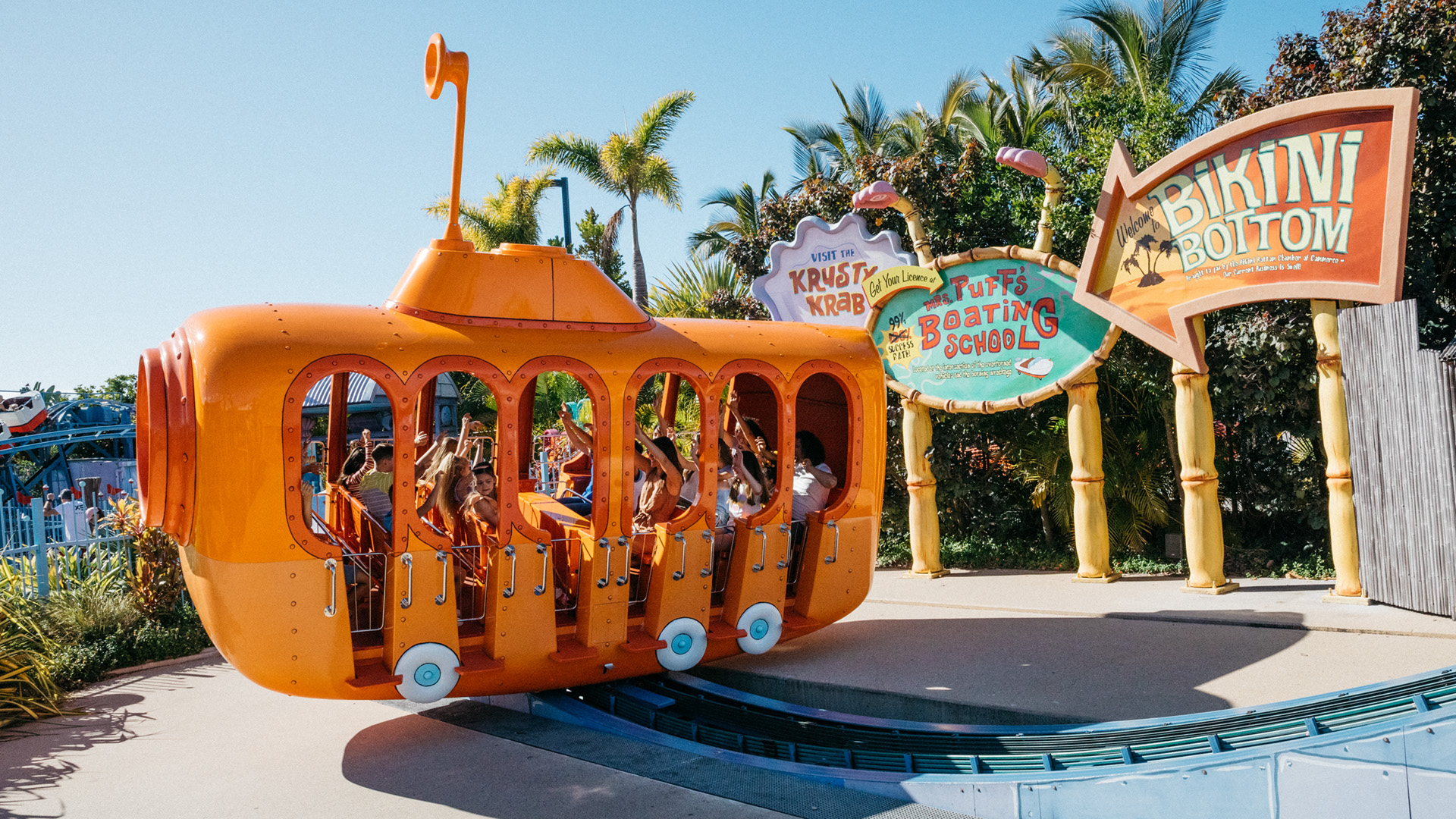 People are seated in an orange amusement ride shaped like a bus with signs for "Krusty Krab" and "Bikini Bottom" in the background surrounded by tropical foliage.