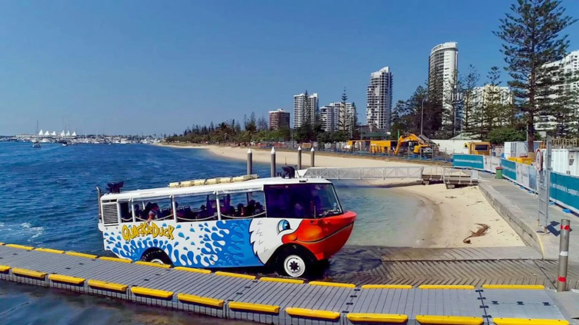 A colorful amphibious bus with a painted smiling face drives from the water onto a dock near a sandy beach, with tall buildings and trees in the background under a clear blue sky.