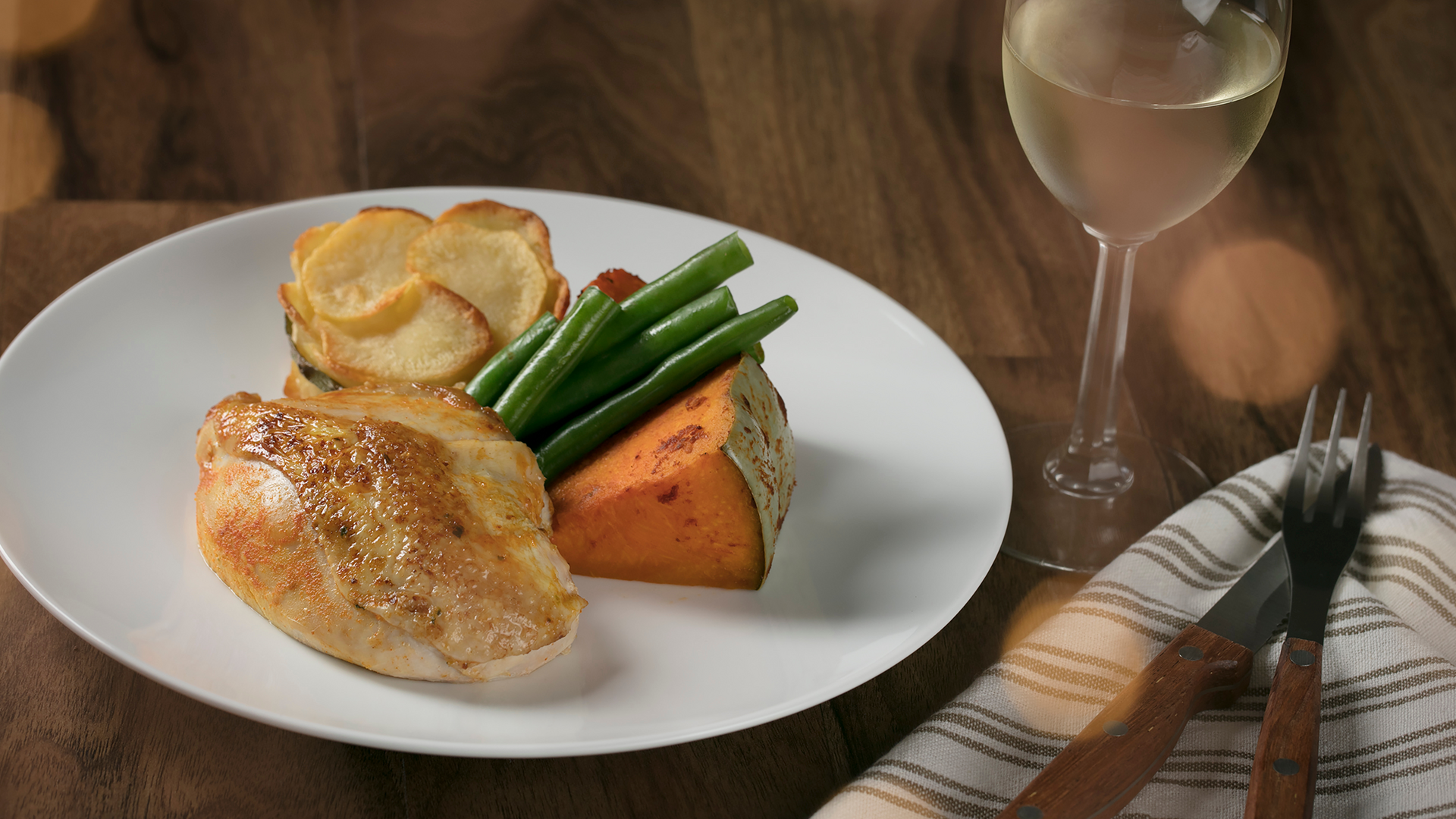 A white plate with roast chicken breast, green beans, a slice of roasted sweet potato, and a potato gratin stack. A glass of white wine and utensils on a striped napkin are beside the plate on a wooden table.