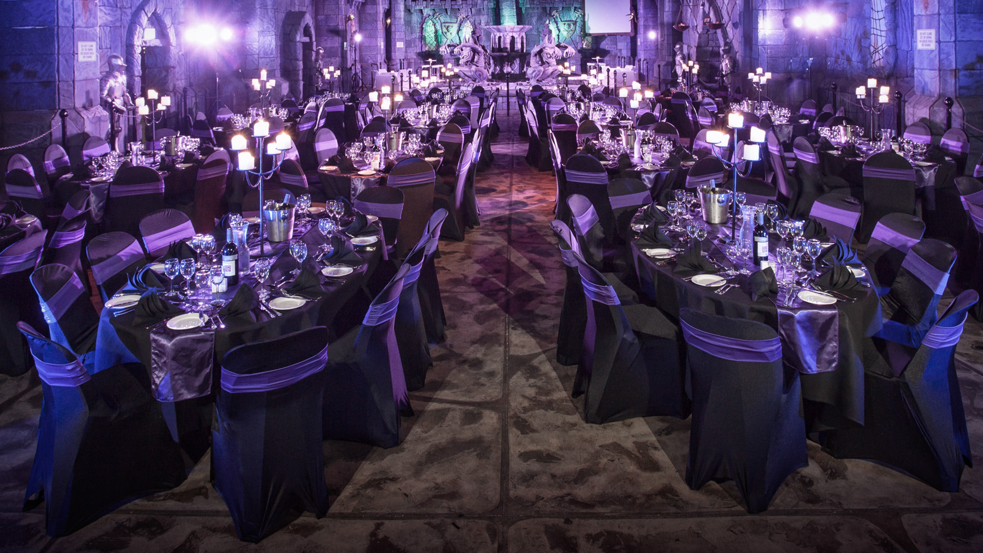A grand banquet hall is decorated with round tables covered in black and purple linens, set with glassware and plates. Candlelit centerpieces create a dramatic, elegant atmosphere with purple lighting illuminating the space.