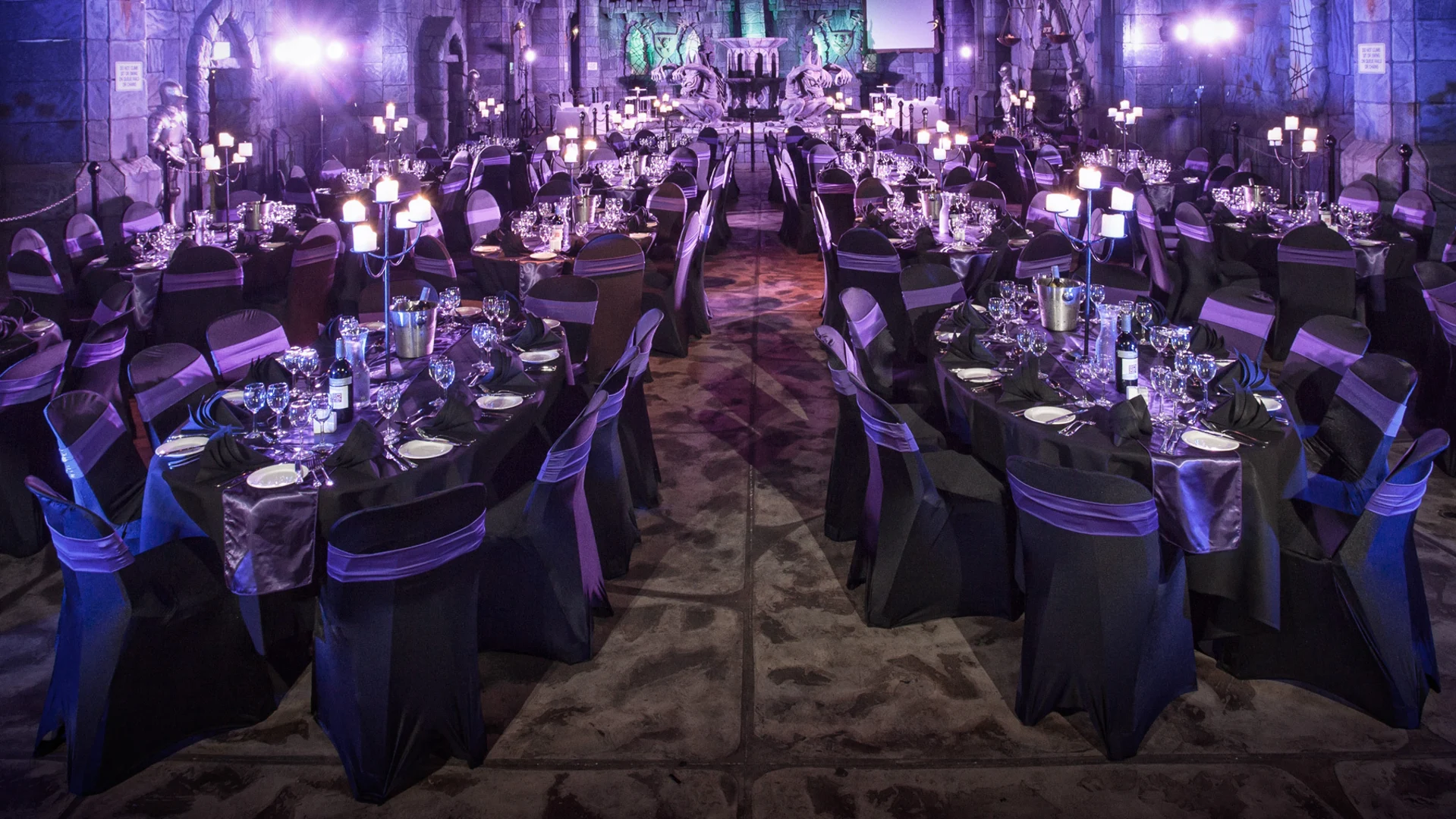 A grand banquet hall is decorated with round tables covered in black and purple linens, set with glassware and plates. Candlelit centerpieces create a dramatic, elegant atmosphere with purple lighting illuminating the space.