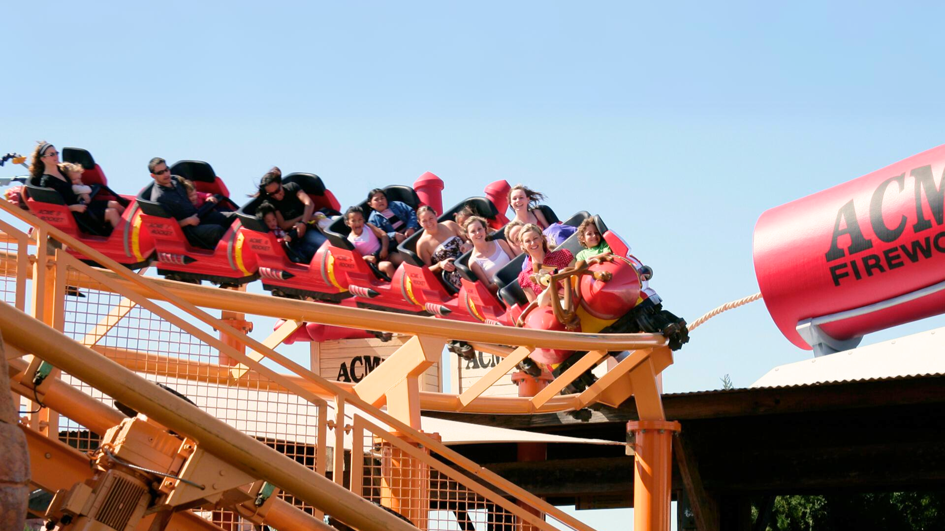 A group of people ride a red and yellow roller coaster with large ACME Fireworks props in the background on a clear sunny day.