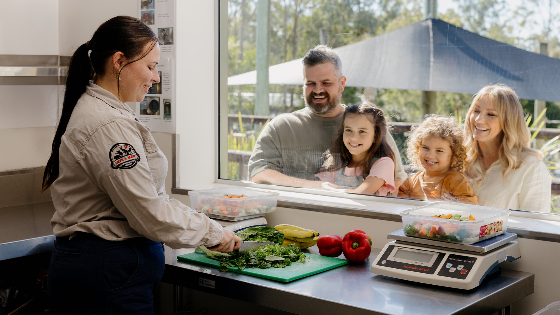A woman in uniform chops vegetables on a counter indoors while a smiling family—two adults and two children—watches her through a large window from outside. Various fresh produce and a scale are on the counter.
