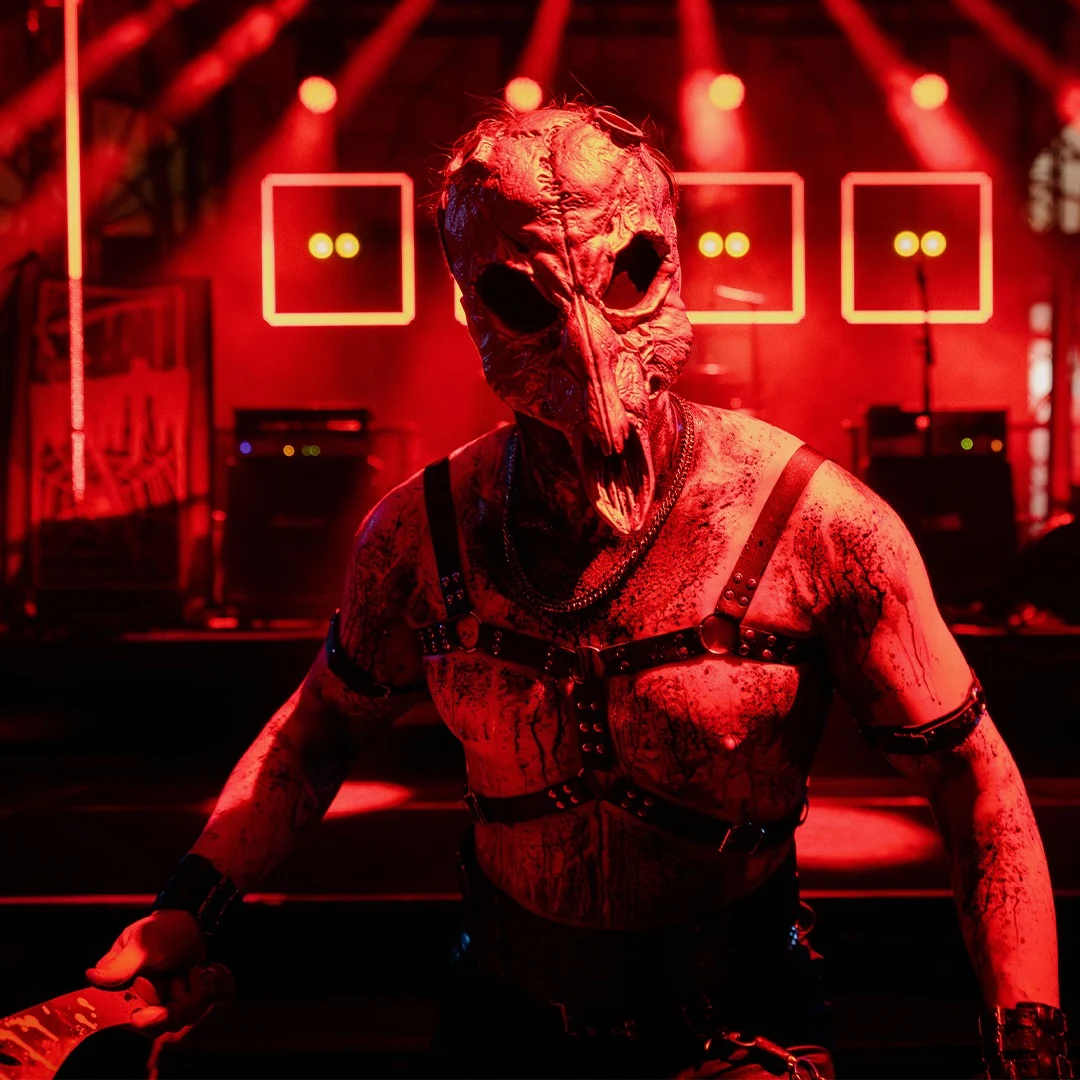 A person wearing a skull mask and leather straps stands under red stage lights in front of a concert setup with speakers and illuminated squares in the background.