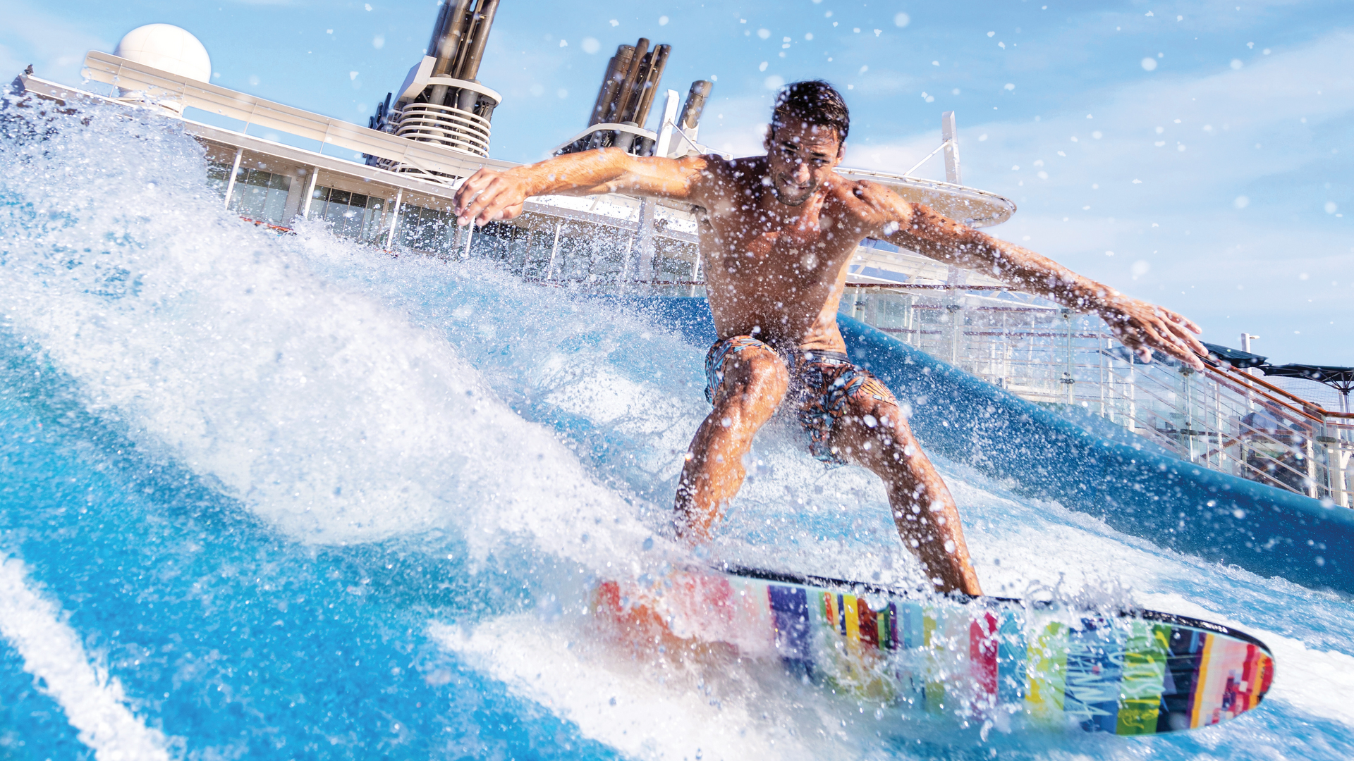 A man in swim trunks rides a colorful surfboard on an artificial wave, with splashing water around him and a cruise ship structure visible in the background under a clear blue sky.