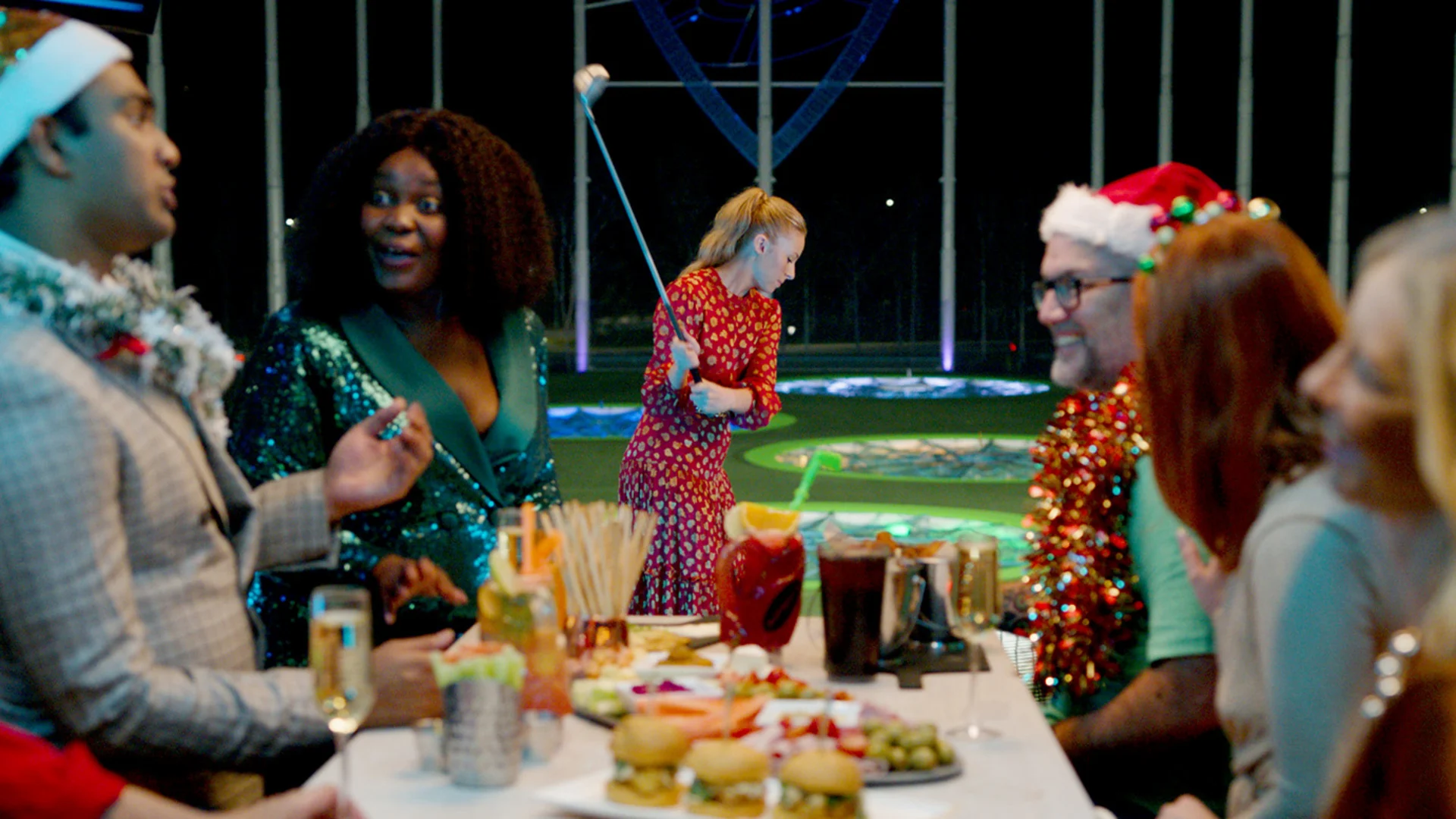 A group of people in festive attire enjoy food and drinks at a decorated table, while a woman in a red dress plays golf in the background. Some wear Santa hats and tinsel, creating a holiday party atmosphere.