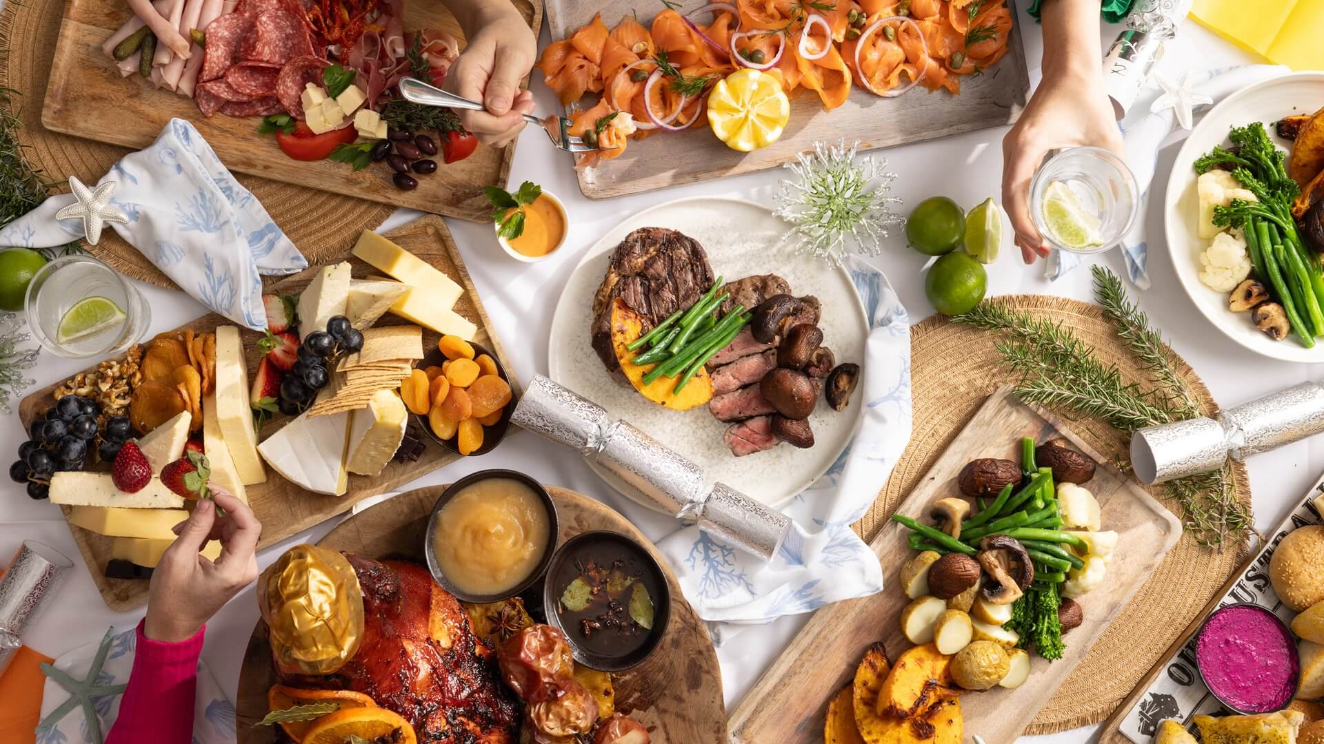 A festive table filled with various dishes including grilled meat, smoked salmon, cheese platter with fruit, roasted vegetables, and drinks, with several hands reaching for food.
