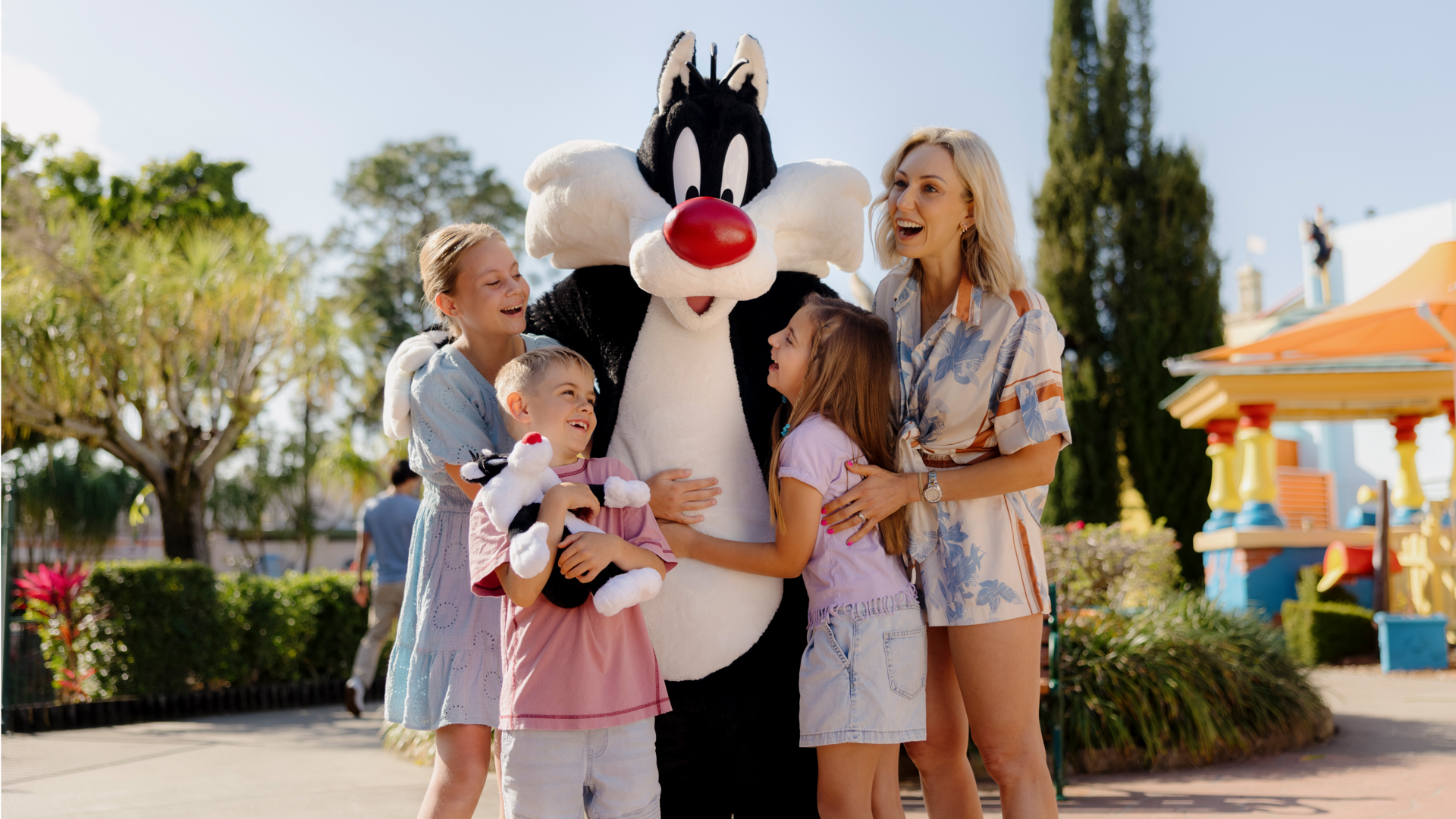 A costumed Sylvester the Cat poses outdoors with three smiling children and a woman, all laughing and embracing the character on a sunny day at an amusement park.