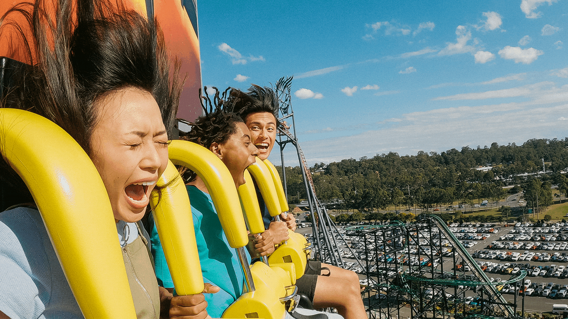 Three people are screaming and holding onto their restraints as they ride a roller coaster with a clear sky and a view of a parking lot and trees in the background.