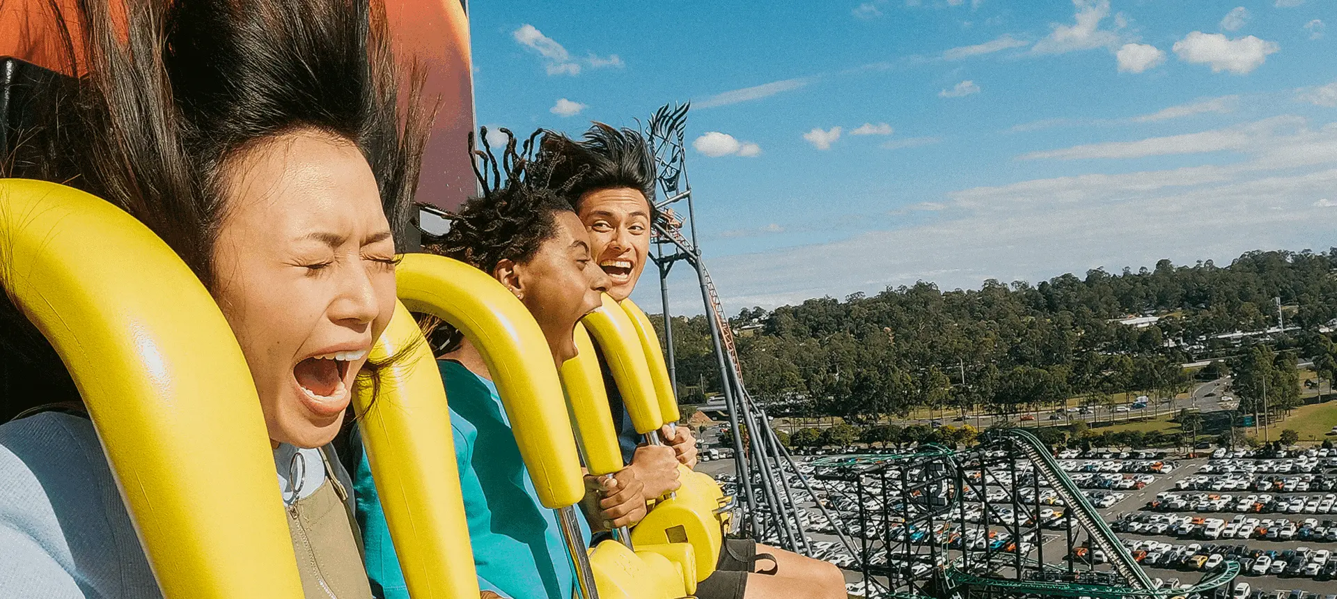 Three people are screaming and holding onto their restraints as they ride a roller coaster with a clear sky and a view of a parking lot and trees in the background.