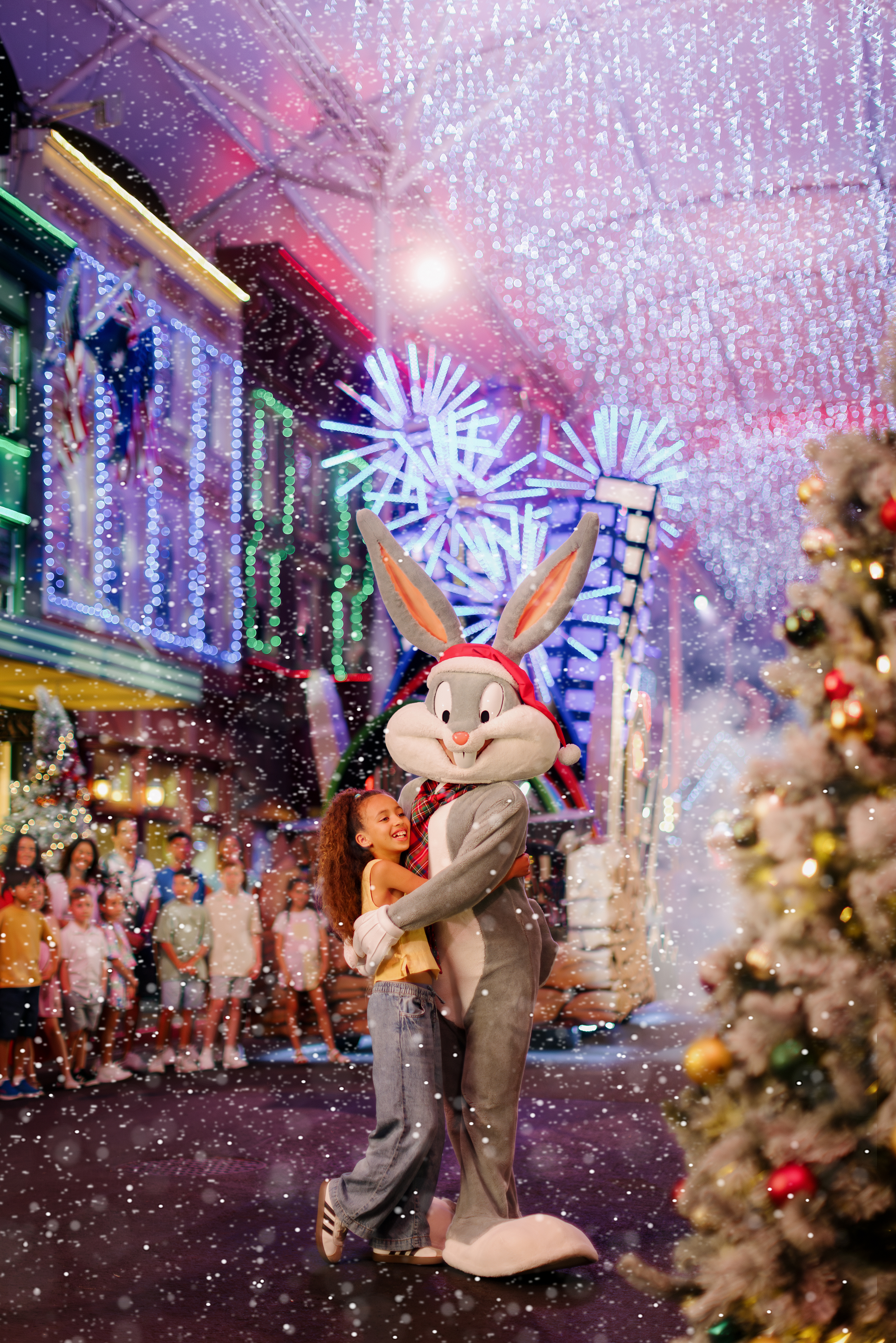 A person hugs a Bugs Bunny mascot in a snowy, festive street filled with colorful lights, holiday decorations, and a Christmas tree, with a crowd of people watching in the background.
