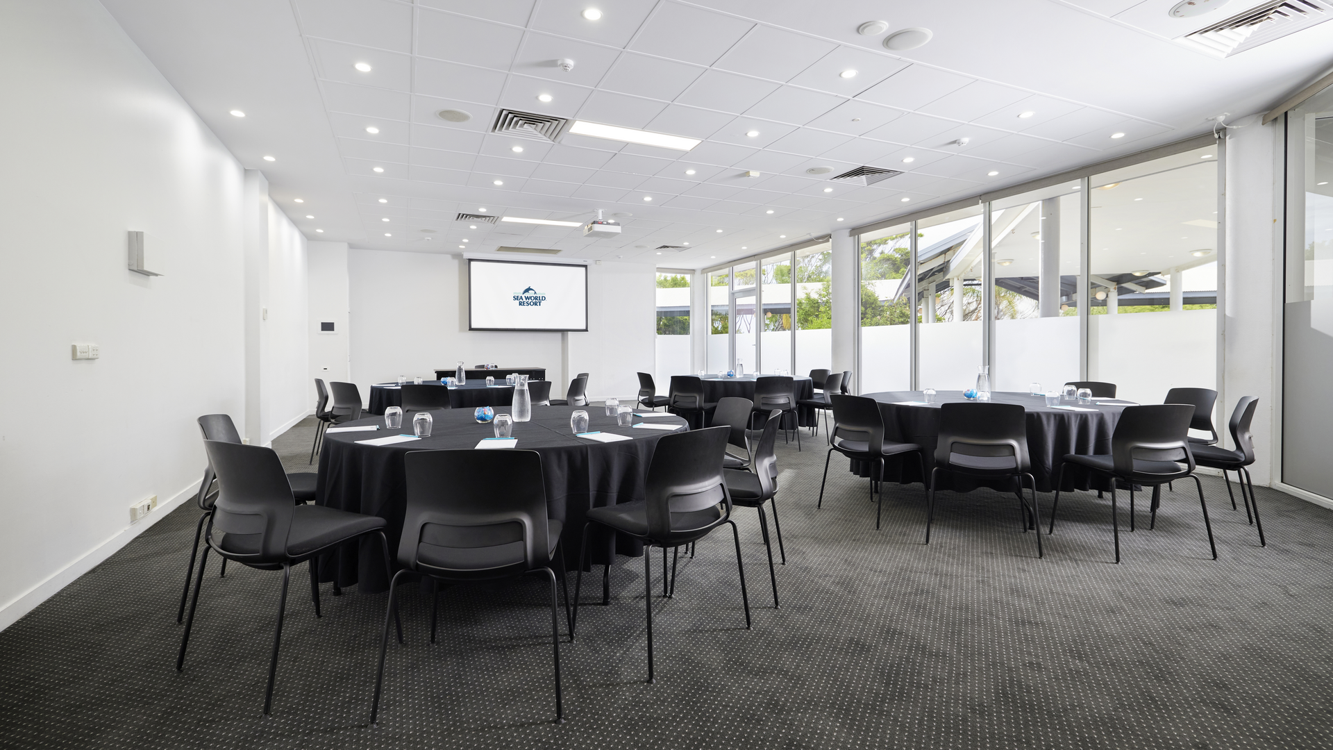 A modern conference room with round tables covered in black tablecloths, surrounded by black chairs. A projection screen is at one end, and large windows line one wall, letting in natural light. The room is empty.