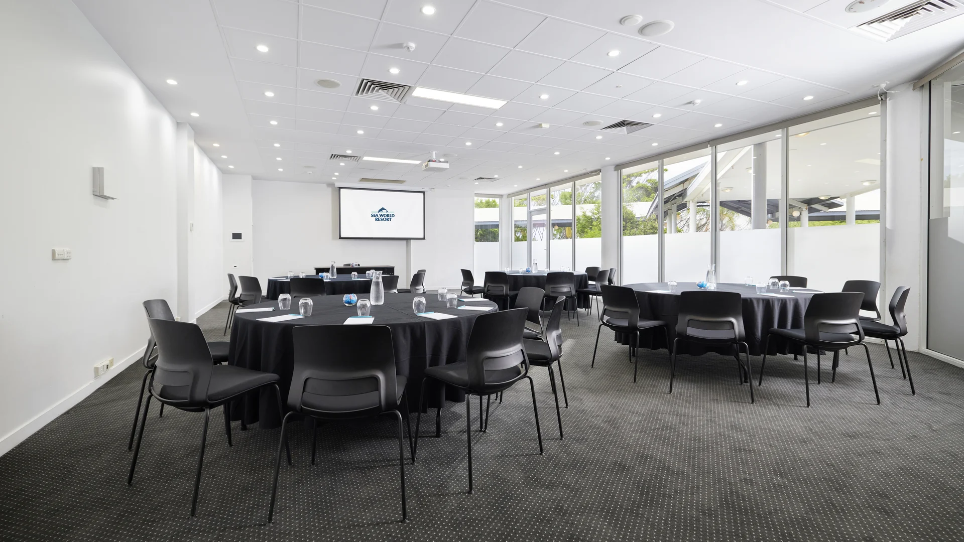 A modern conference room with round tables covered in black tablecloths, surrounded by black chairs. A projection screen is at one end, and large windows line one wall, letting in natural light. The room is empty.