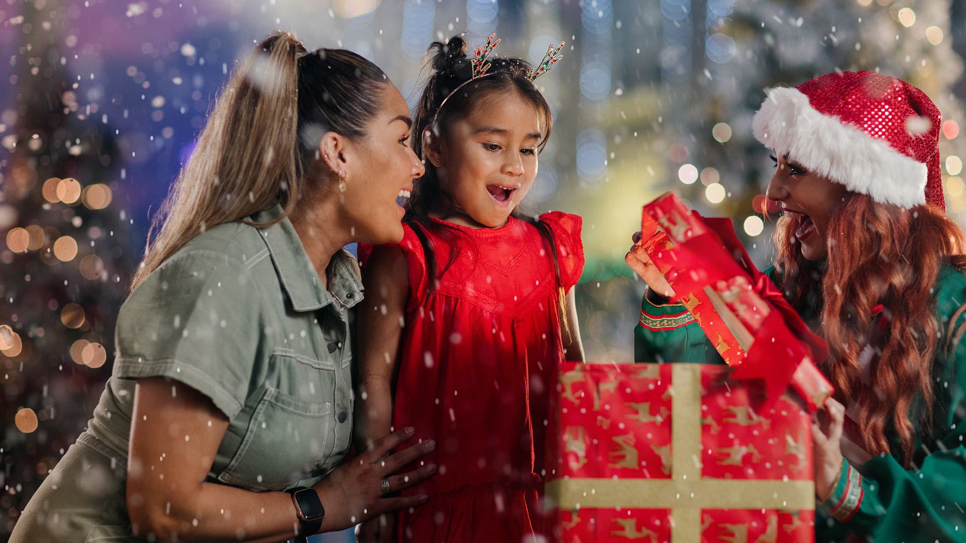 Three people celebrate Christmas, standing in falling snow. A child in a red dress looks excitedly at a large present held by a woman in a Santa hat, while another woman smiles beside her. Festive lights glow in the background.