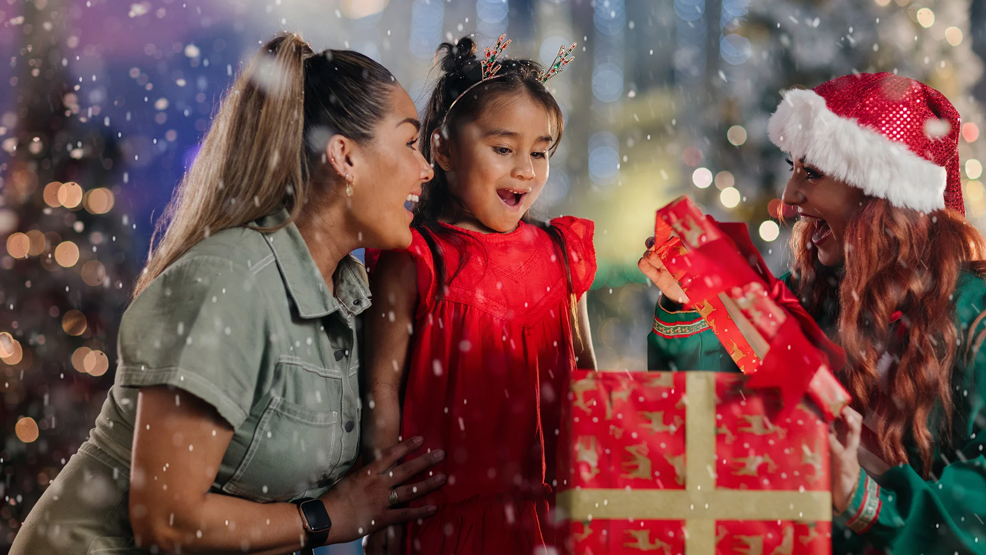 Three people celebrate Christmas, standing in falling snow. A child in a red dress looks excitedly at a large present held by a woman in a Santa hat, while another woman smiles beside her. Festive lights glow in the background.