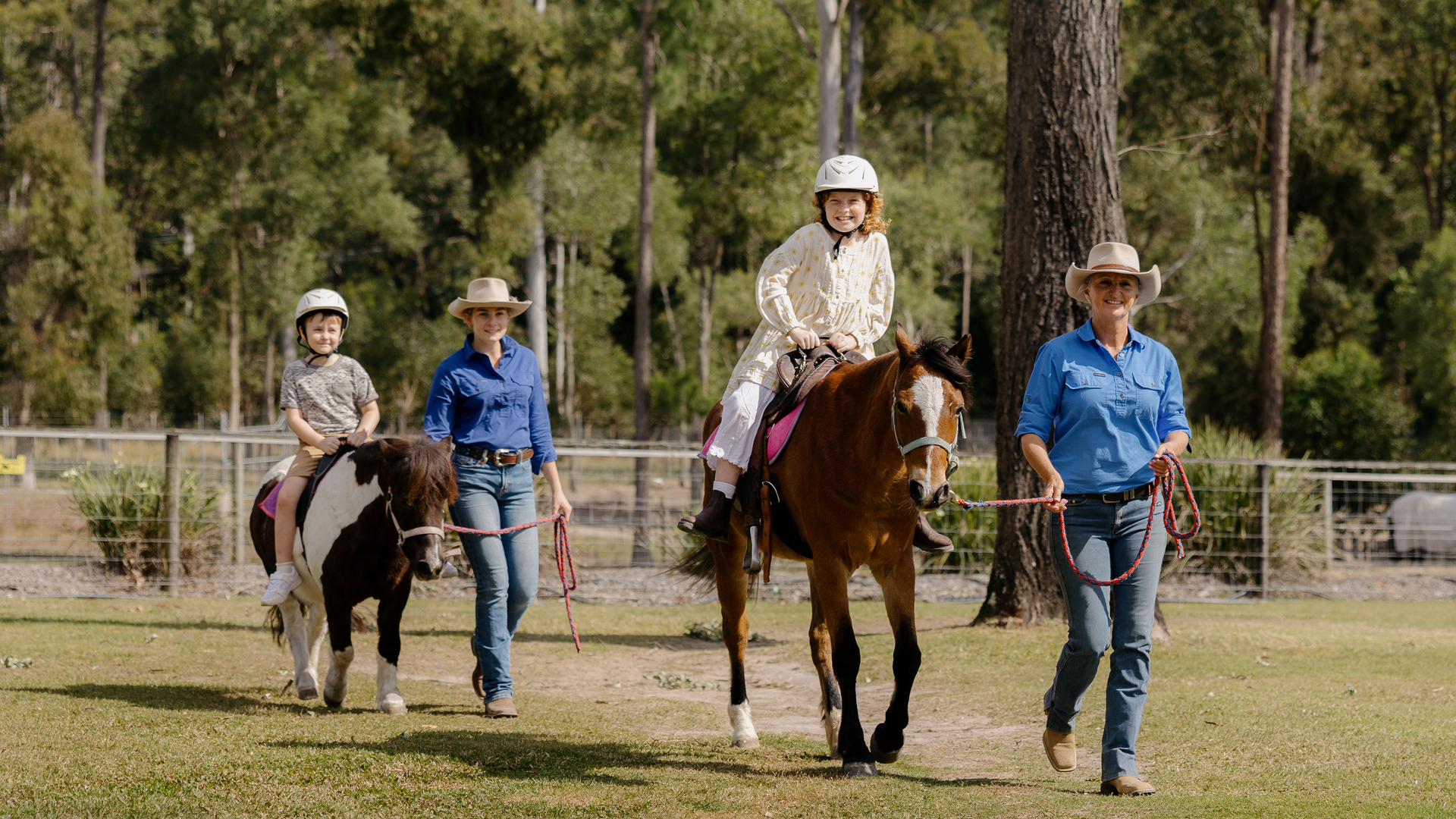 Two children with helmets ride ponies, each led by an adult in blue shirts and hats, walking on grass with trees and a fence in the background.