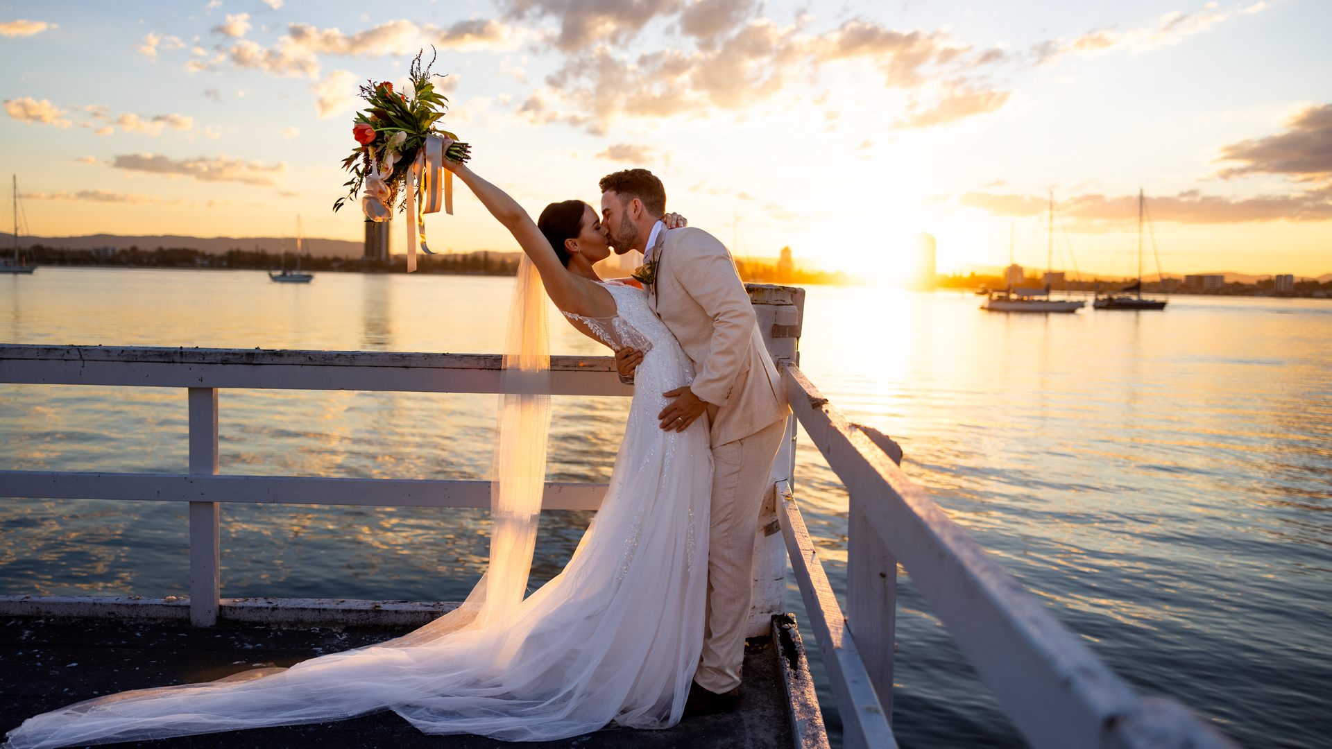 A bride and groom kiss on a dock at sunset, with the bride holding a bouquet and her veil flowing behind her. Sailboats and a golden sky are visible in the background over calm water.