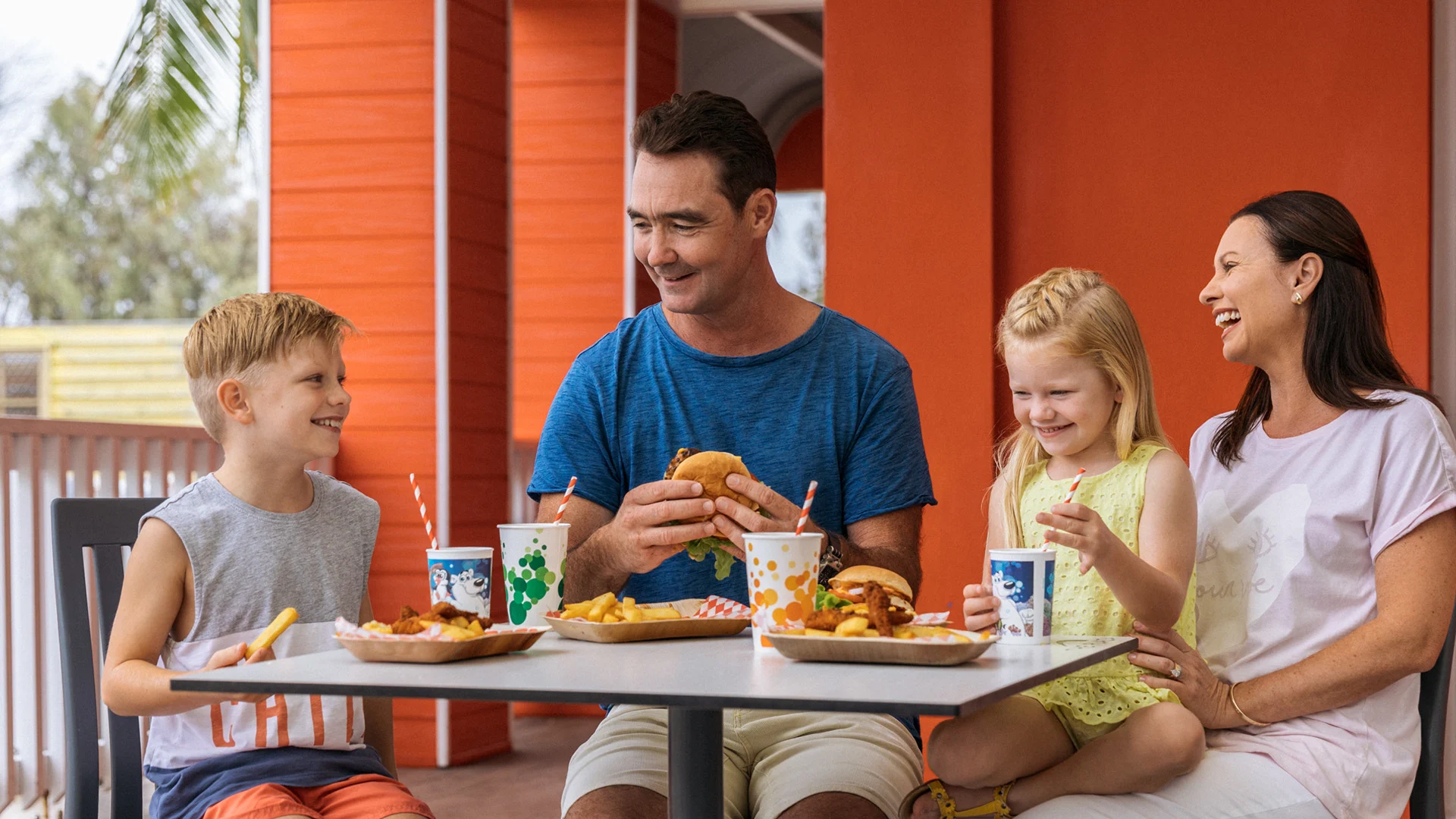 A family of four sits at an outdoor table eating burgers, fries, and drinking from cups with straws, smiling and enjoying their meal together.