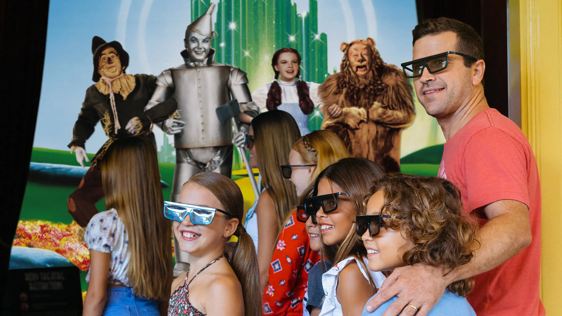 A man and four children wearing 3D glasses stand in front of a "Wizard of Oz" backdrop featuring characters from the movie.