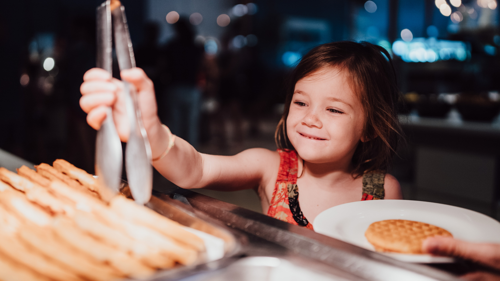 A young girl smiles while using tongs to pick up food from a tray at a buffet. She holds a plate with a waffle in her other hand. The background is softly lit and out of focus.