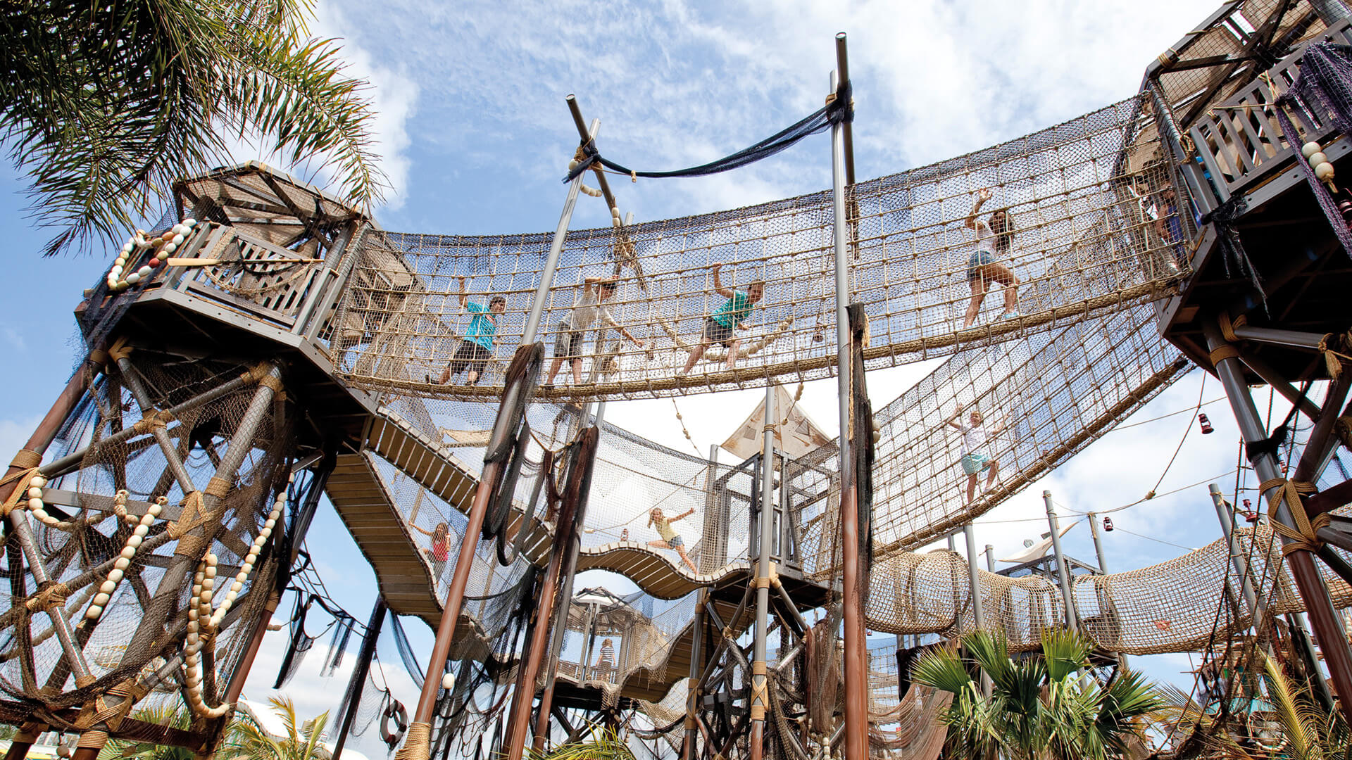 Children and adults walk and climb on elevated rope bridges and wooden platforms in an outdoor adventure playground under a partly cloudy sky.