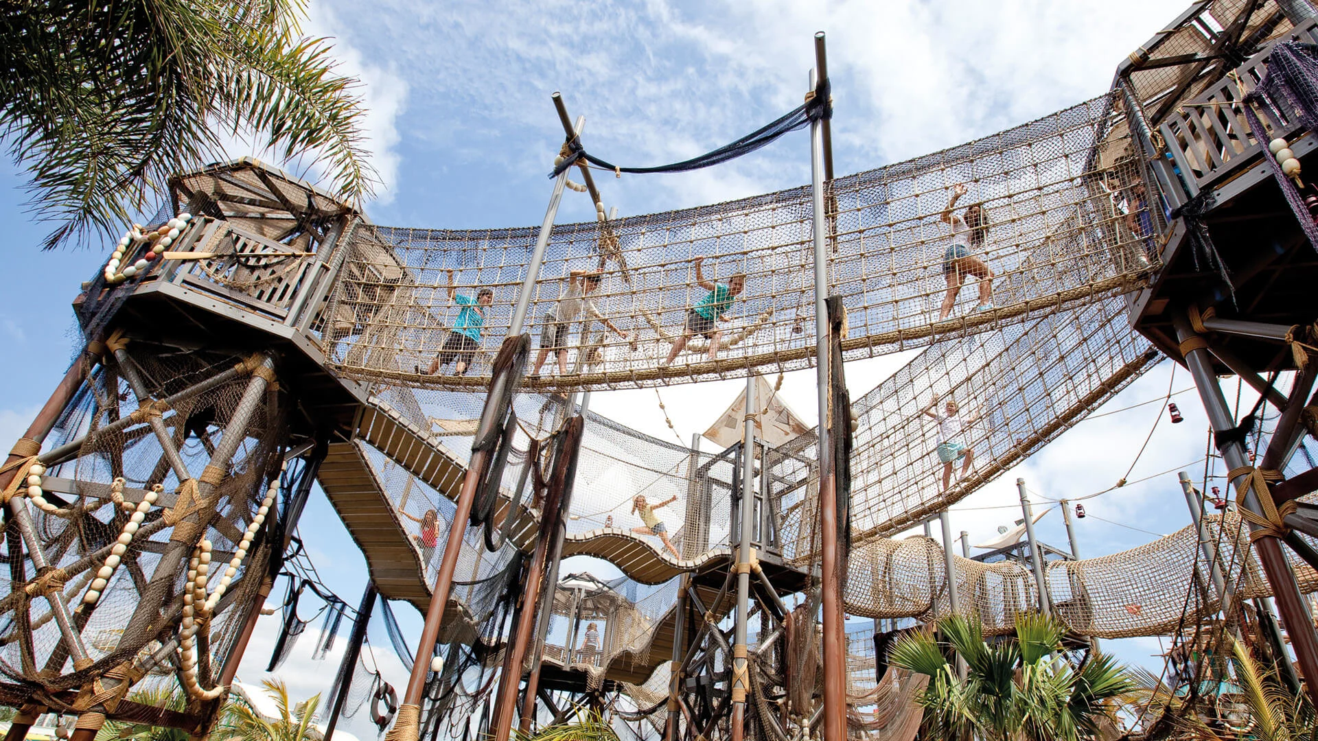 Children and adults walk and climb on elevated rope bridges and wooden platforms in an outdoor adventure playground under a partly cloudy sky.