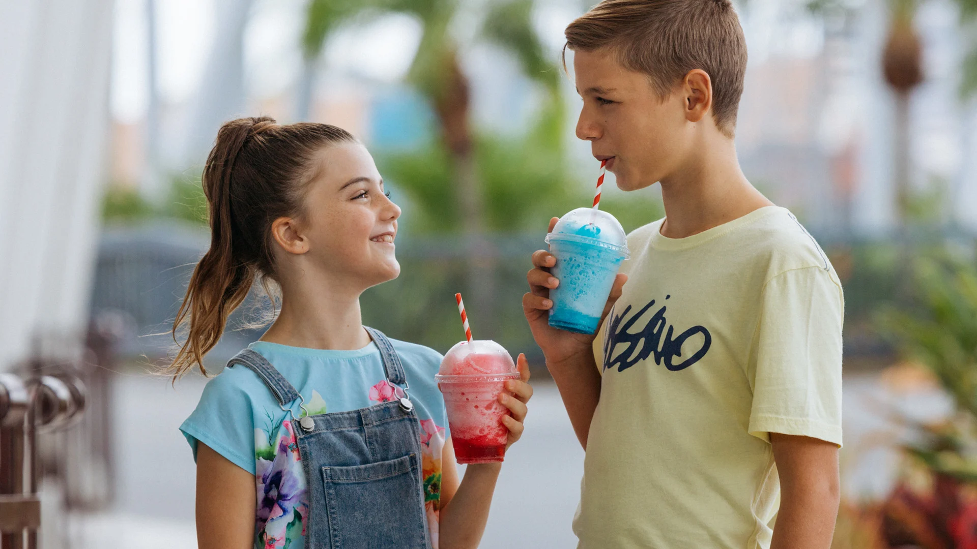 A girl and a boy stand outside drinking colorful slushies with striped straws, smiling at each other.